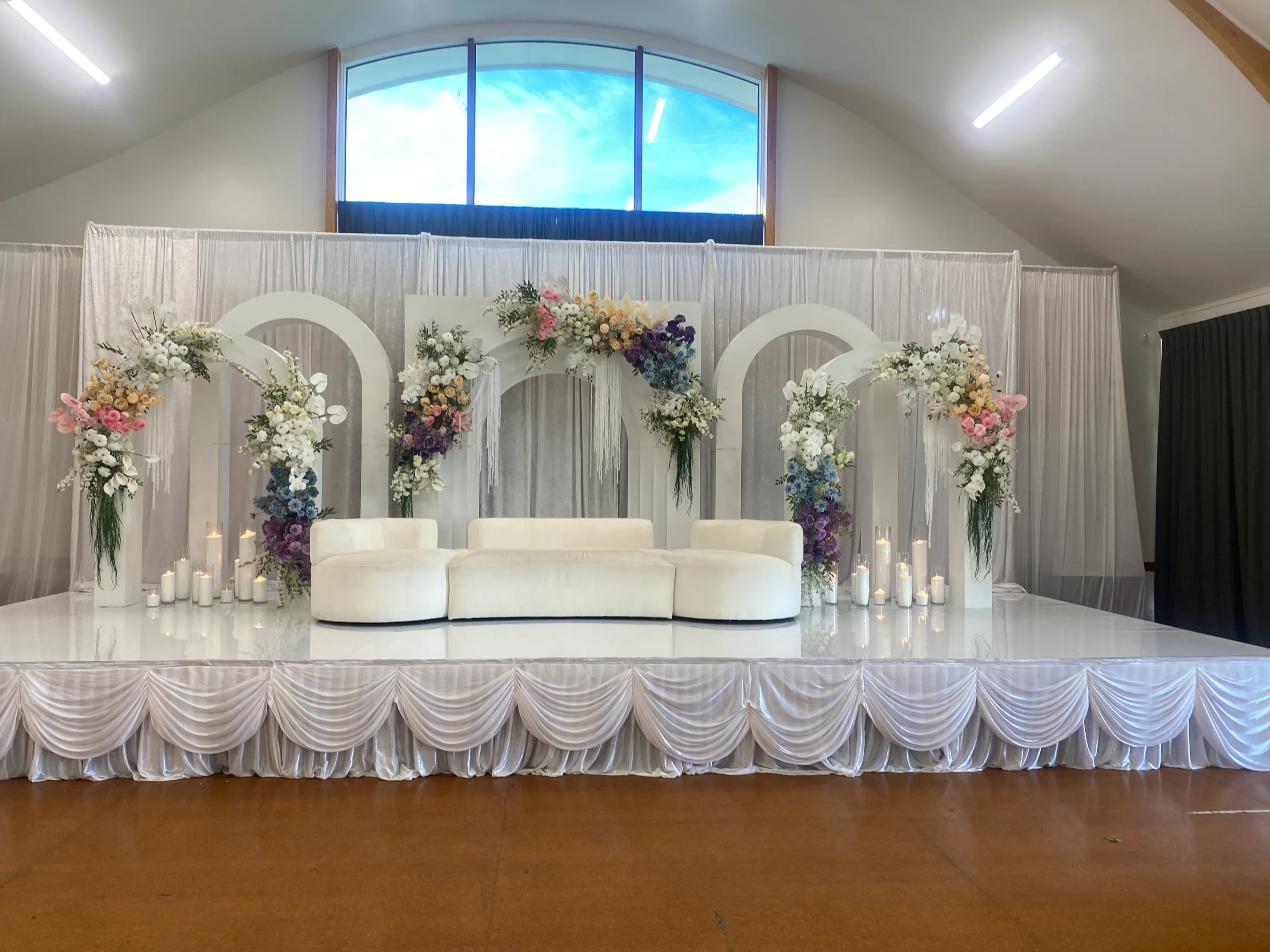 Stage styling by Decoration X: Front view of a glossy white stage with satin swag skirting, a curved cream sofa, four white arches with pastel and blue florals, and pillar candles in glass.