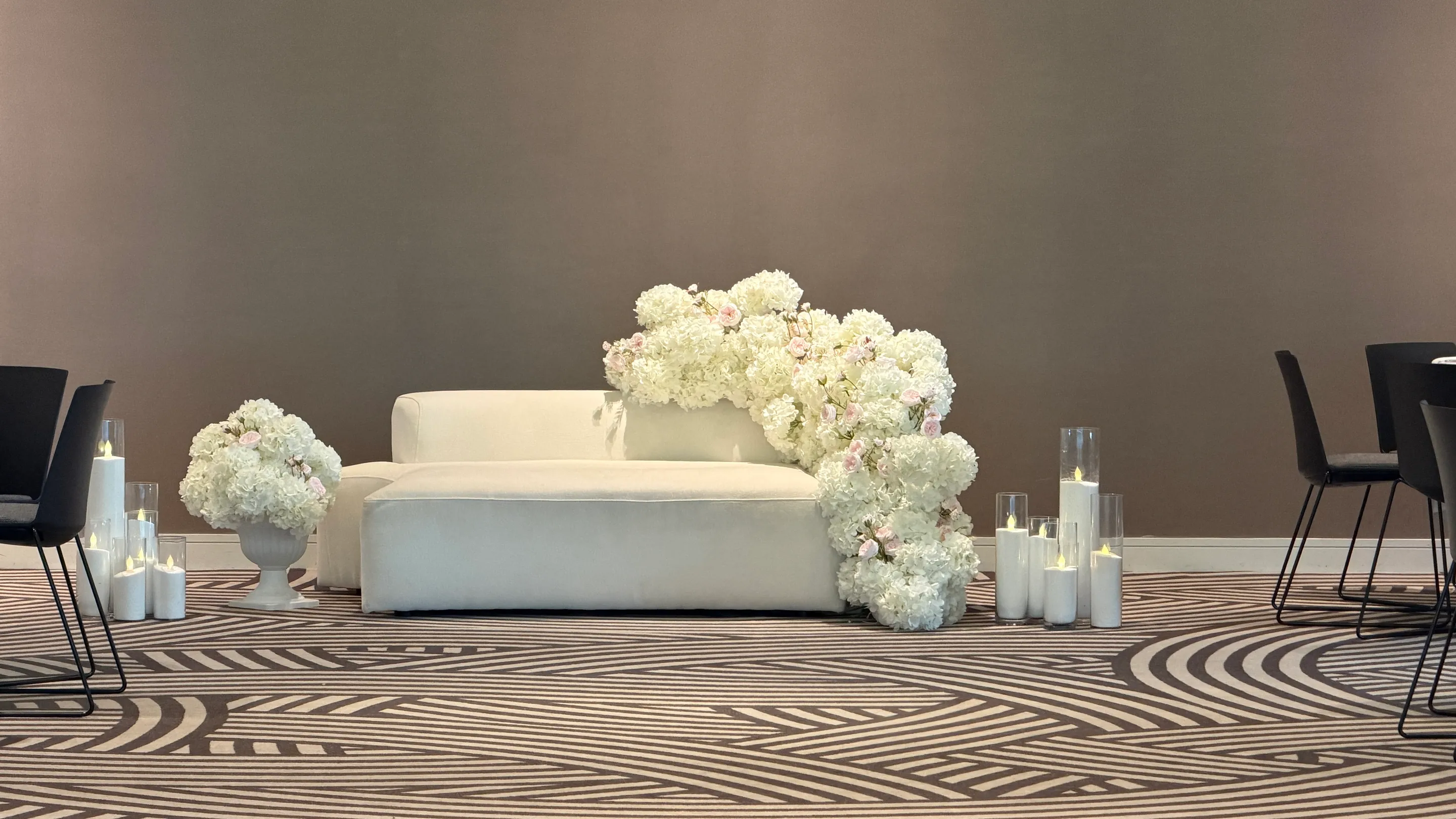 Stage styling by Decoration X: White modular sofa with a cascade of white hydrangeas and blush roses, a hydrangea urn and pillar candles in glass cylinders on a striped patterned carpet.