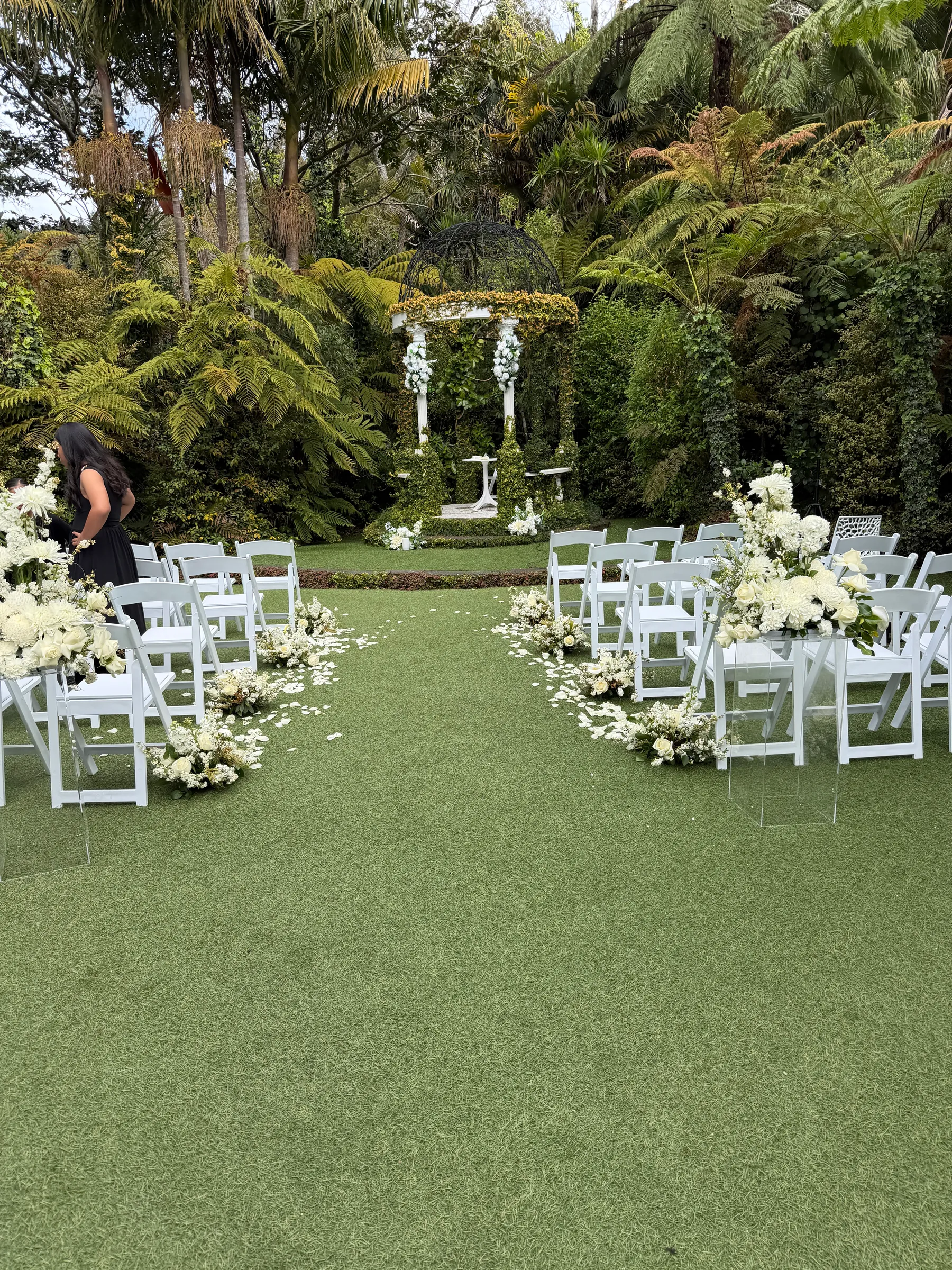 Ceremony styling by Decoration X: Lawn ceremony with white folding chairs either side of a petal-strewn aisle of white floral clusters, leading to a gazebo dressed in white florals among tree ferns.