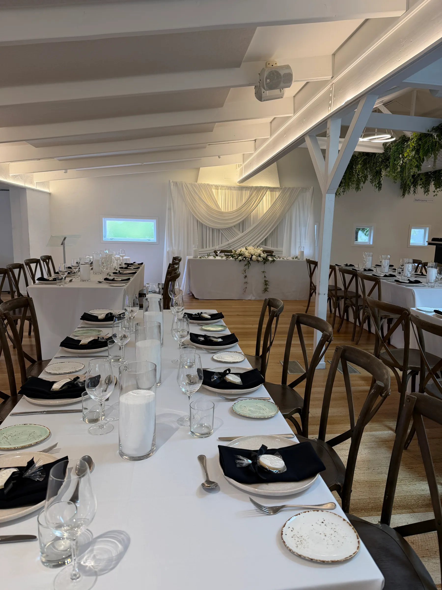 Reception styling by Decoration X: Long tables in white linen set with black napkins, speckled plates, glassware and pillar candles under white beams, a draped head table at the far end.