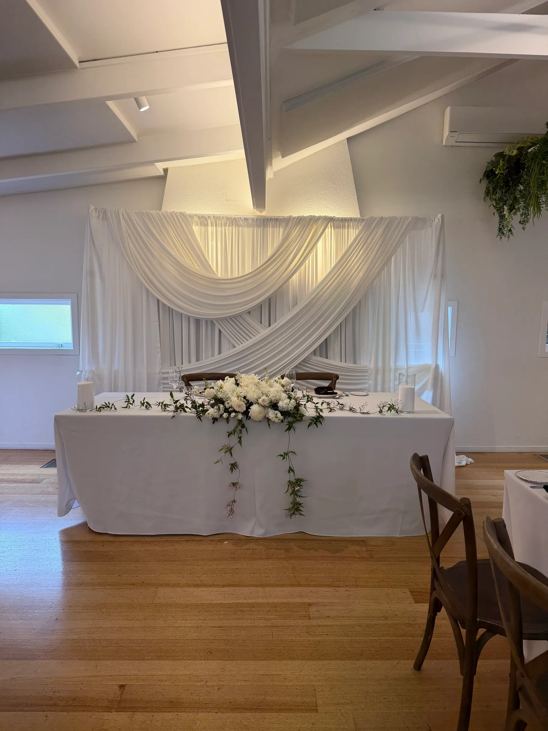 Stage styling by Decoration X: Head table in white linen with a garland of white roses and trailing greenery, crossed white drapes on a backdrop behind, wooden cross-back chairs nearby.