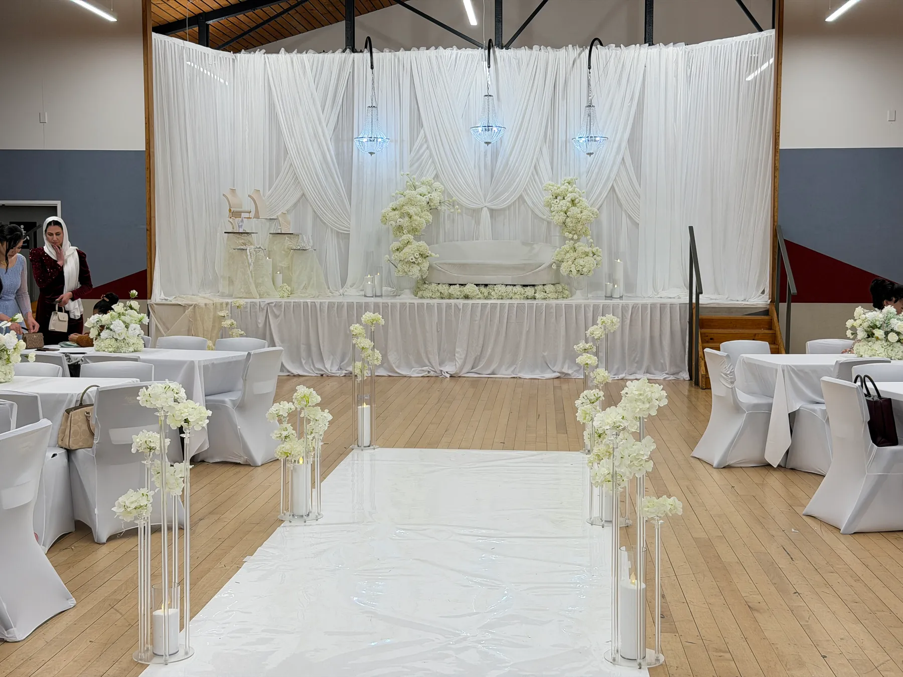 Ceremony styling by Decoration X: White runner aisle lined with tall acrylic stands of white blooms and candles, leading to a white-draped stage with a sofa and chandeliers, guest tables either side.