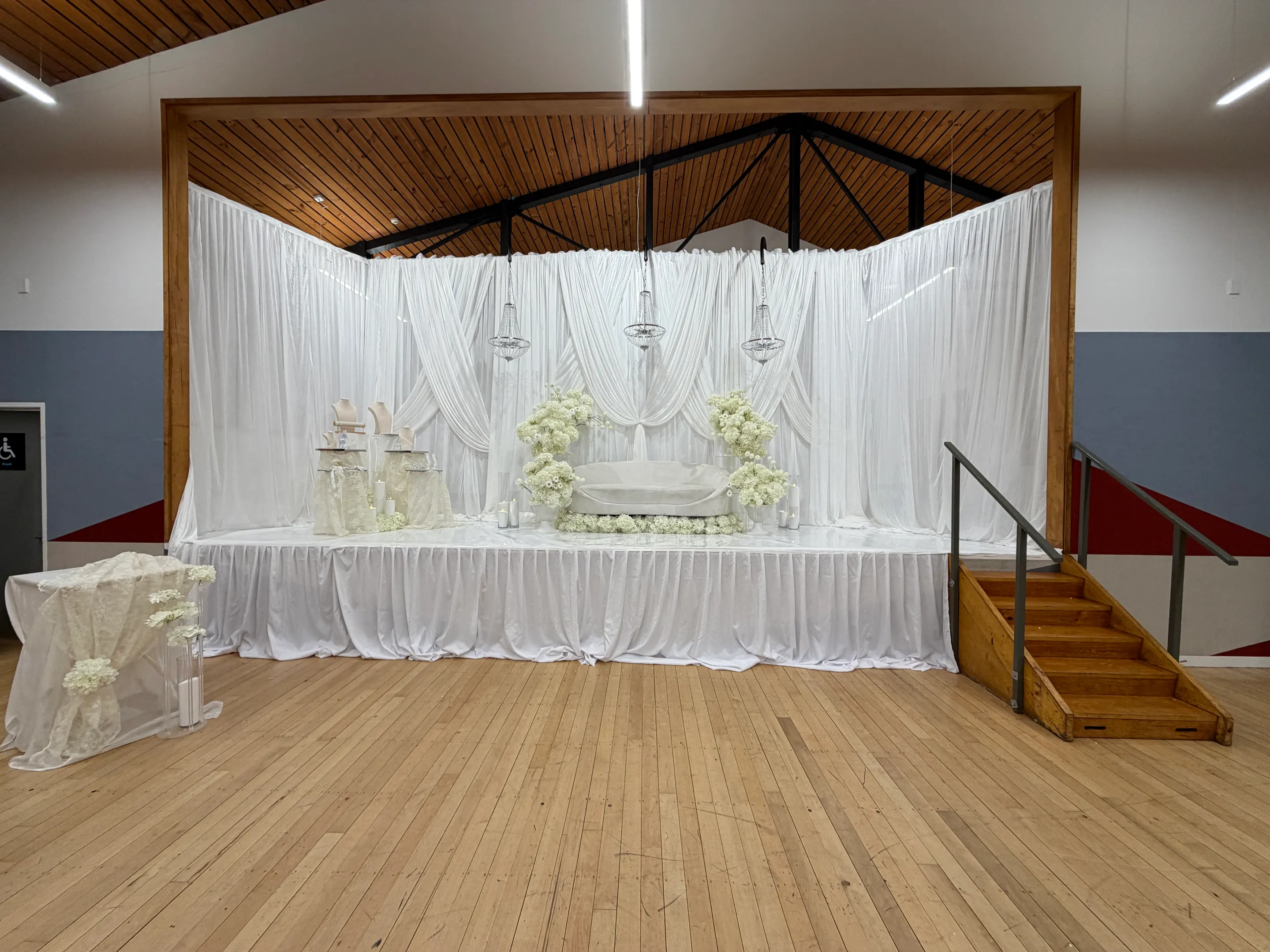 Stage styling by Decoration X: Raised stage skirted and draped in white, a white sofa between white floral columns, jewellery display busts and three crystal chandeliers, wooden steps at the side.