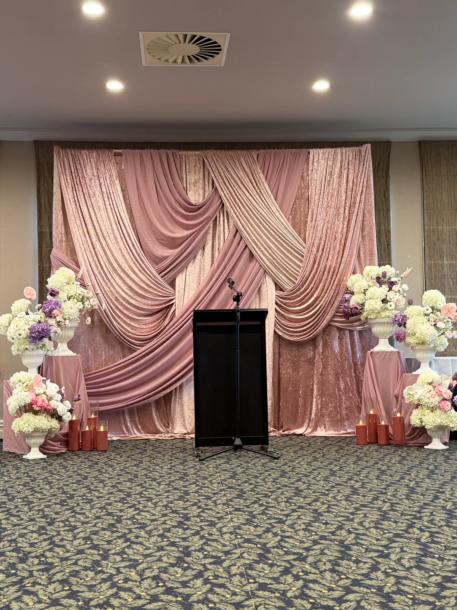 Corporate styling by Decoration X: Crossed swags of blush and dusky pink velvet behind a black lectern and microphone, white and lilac hydrangeas in white urns on draped plinths with rose-gold candles.