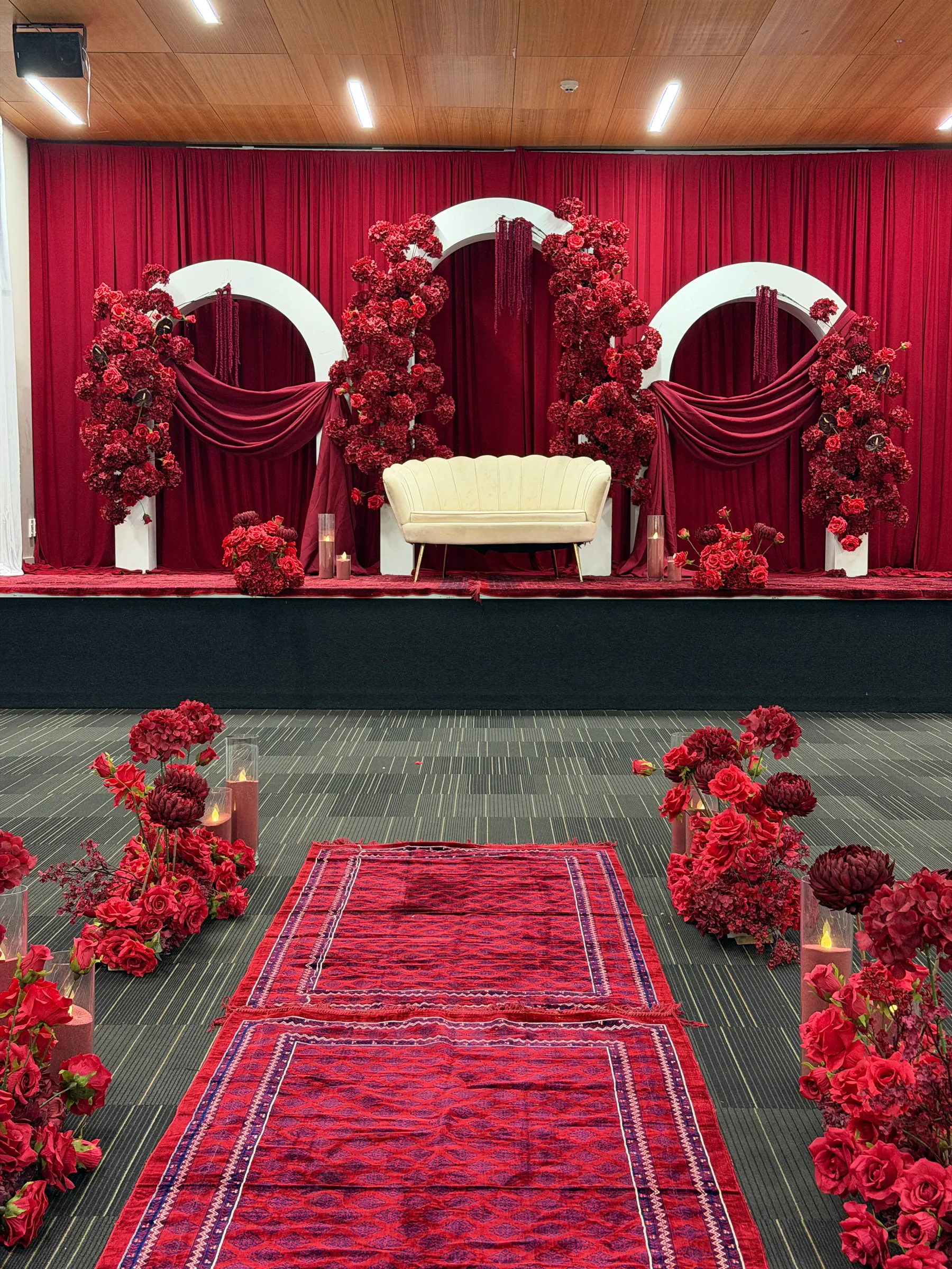 Ceremony styling by Decoration X: Red rug aisle lined with red roses and candles in glass, leading to a stage of white arches, burgundy swags and red rose columns around a cream loveseat.