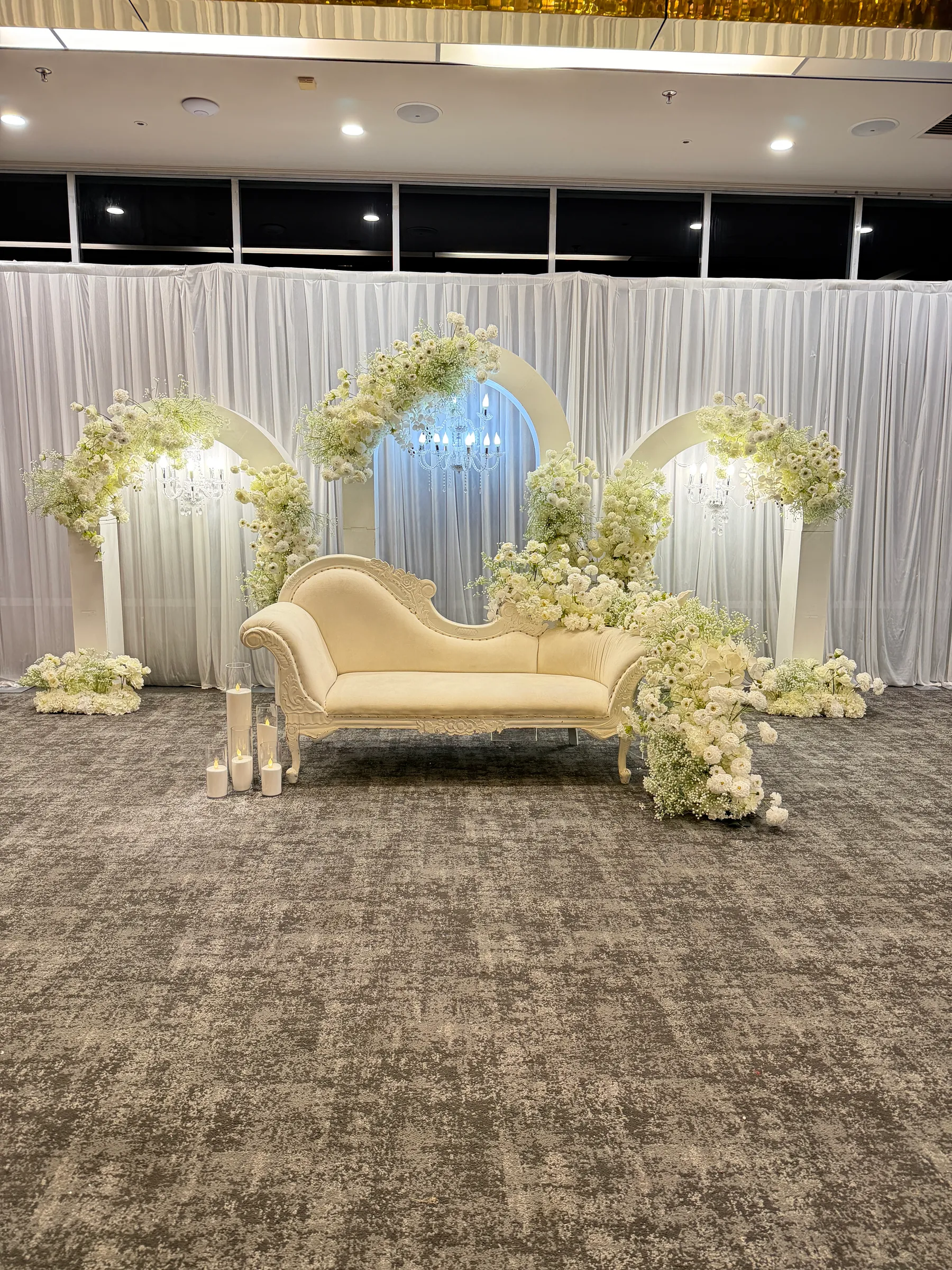 Stage styling by Decoration X: Cream carved chaise before three white arches crowned with white blooms and gypsophila, small chandeliers within, sheer white drapes and pillar candles in glass.
