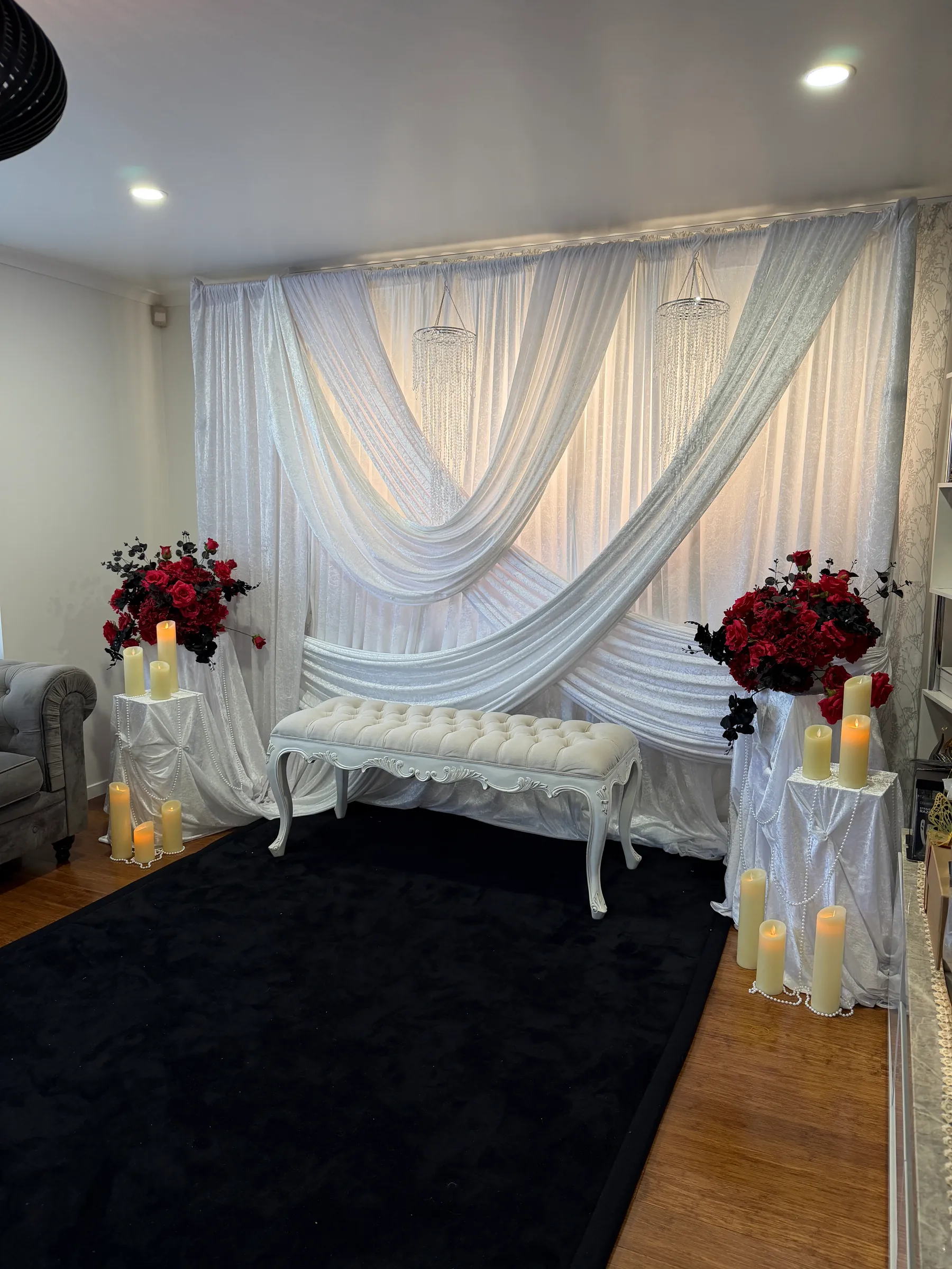 Stage styling by Decoration X: White velvet drapes swagged across a backdrop with two crystal chandeliers, a tufted white bench, red roses on draped plinths and pillar candles on a black rug.