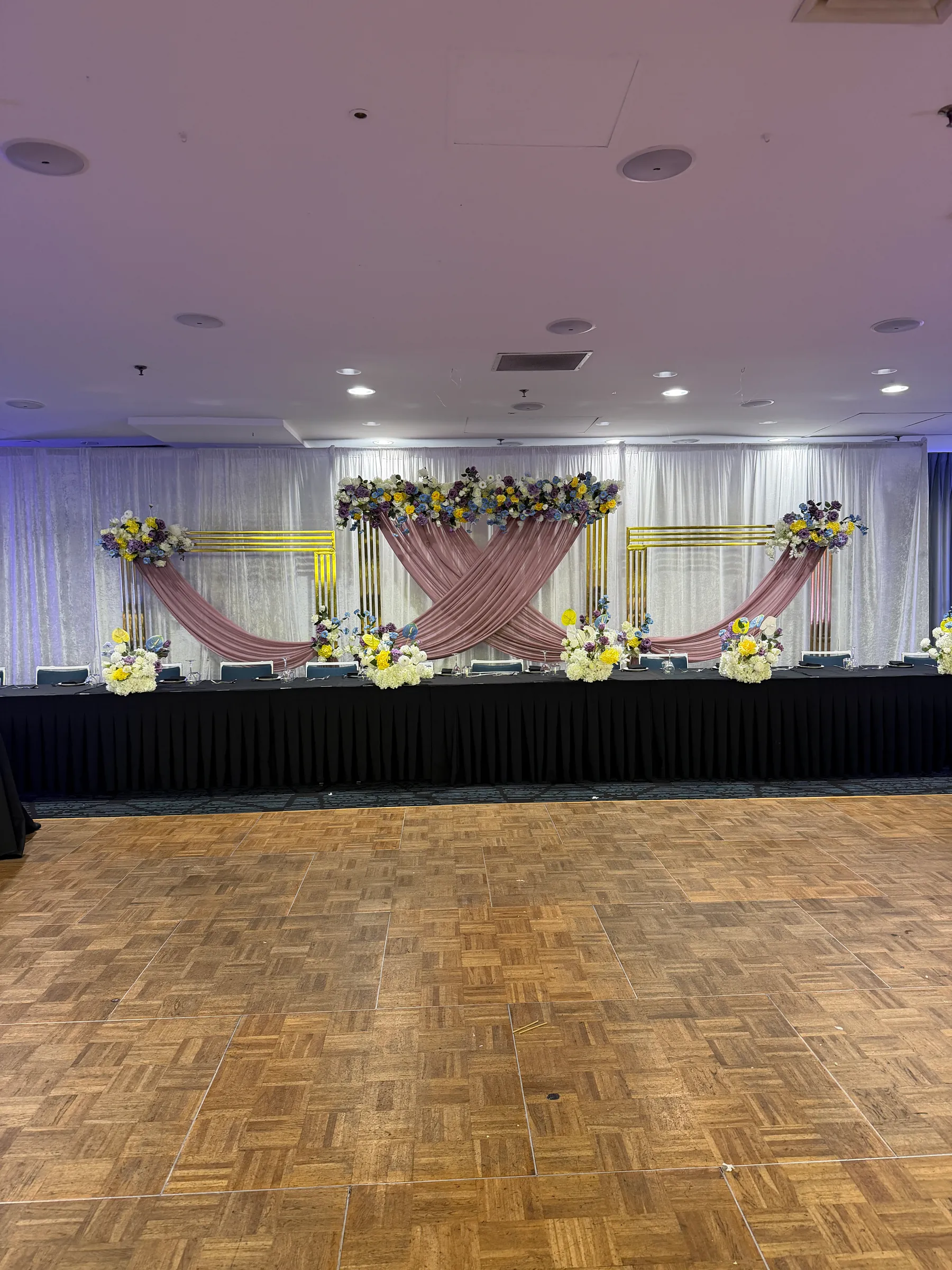 Stage styling by Decoration X: Head table skirted in black before gold frames swagged in dusty pink, mixed florals in purple, yellow, blue and white, parquet dance floor in front.