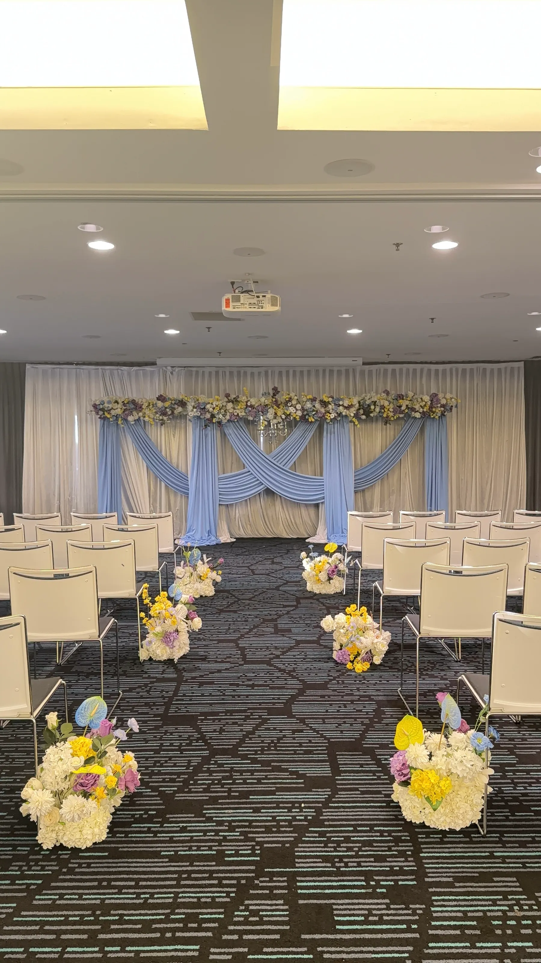 Ceremony styling by Decoration X: Ceremony aisle between white chairs, posies of white hydrangea, yellow and purple blooms and blue anthurium, toward a backdrop of crossed blue drapes.