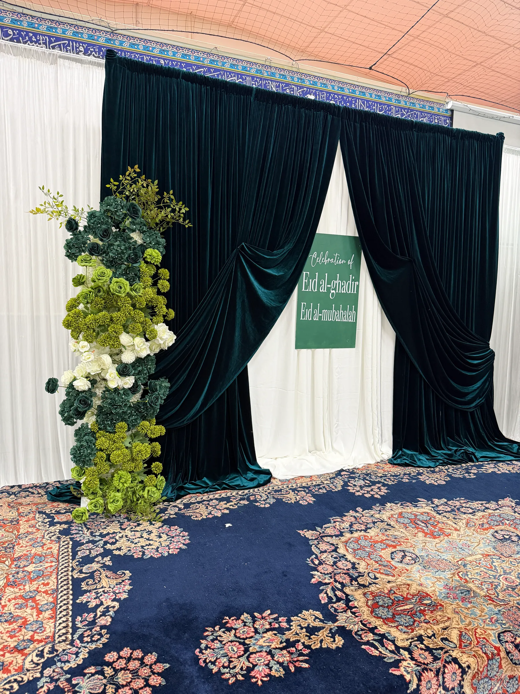 Cultural styling by Decoration X: Teal velvet drapes parted over white, a green Eid sign at centre, a column of green and white roses and hydrangea, Persian rug in navy and red.