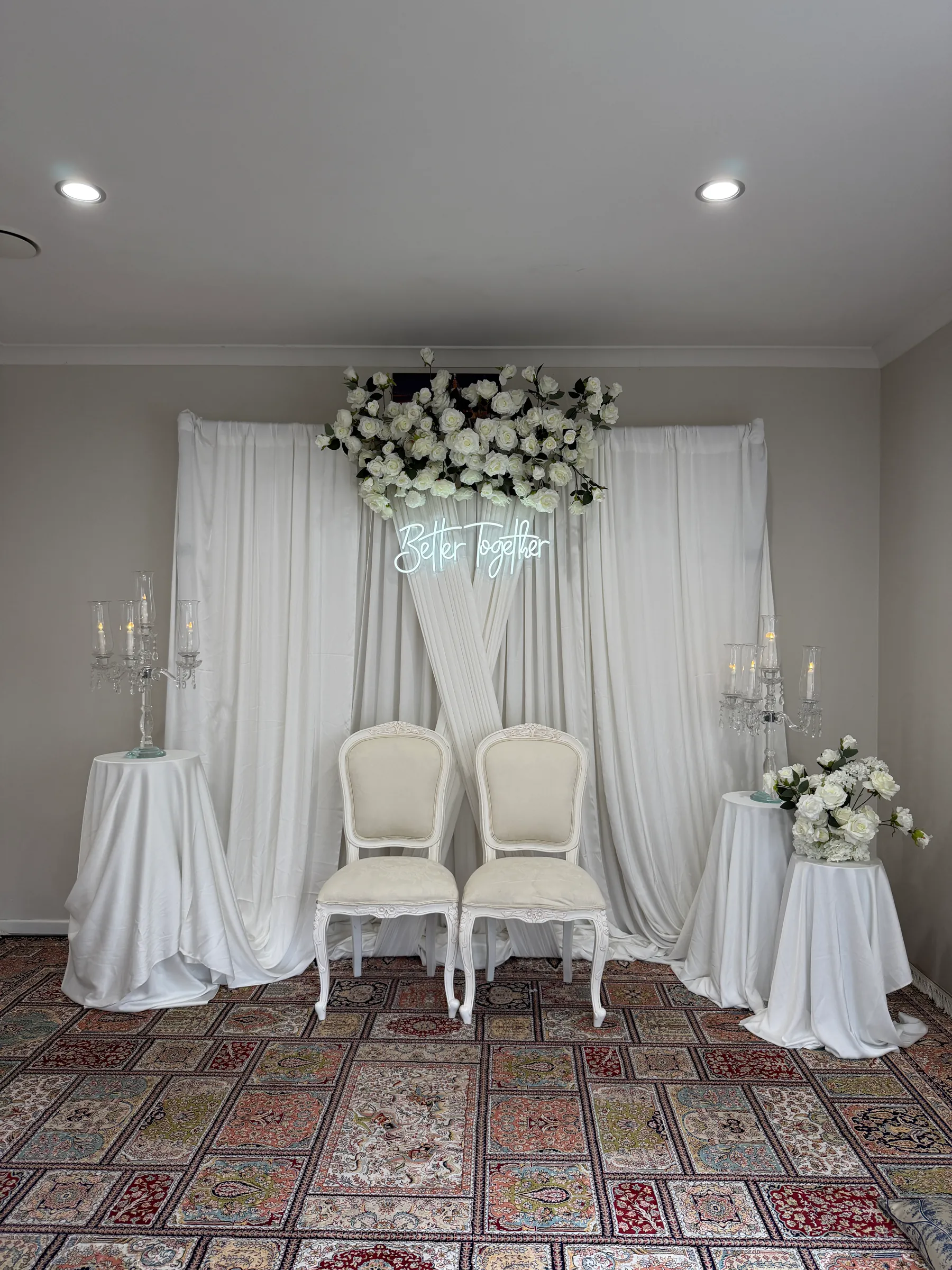 Stage styling by Decoration X: Two carved white chairs before crossed ivory drapes, a white rose swag and neon script sign, crystal candelabra on draped side tables, a patterned rug.