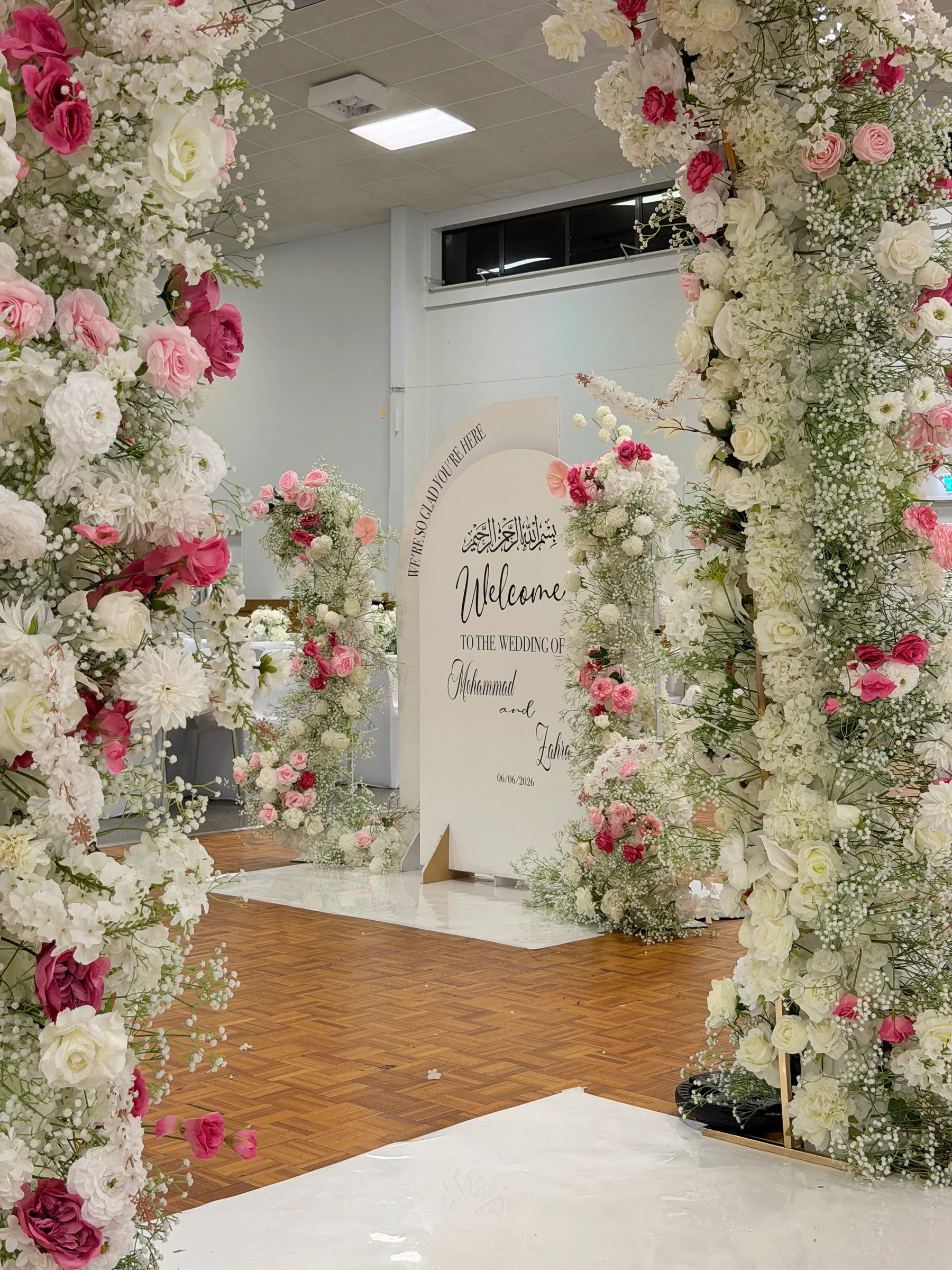 Signage styling by Decoration X: Welcome board with Arabic calligraphy seen through a pink and white rose and gypsophila arch, more floral columns framing it on a parquet floor.