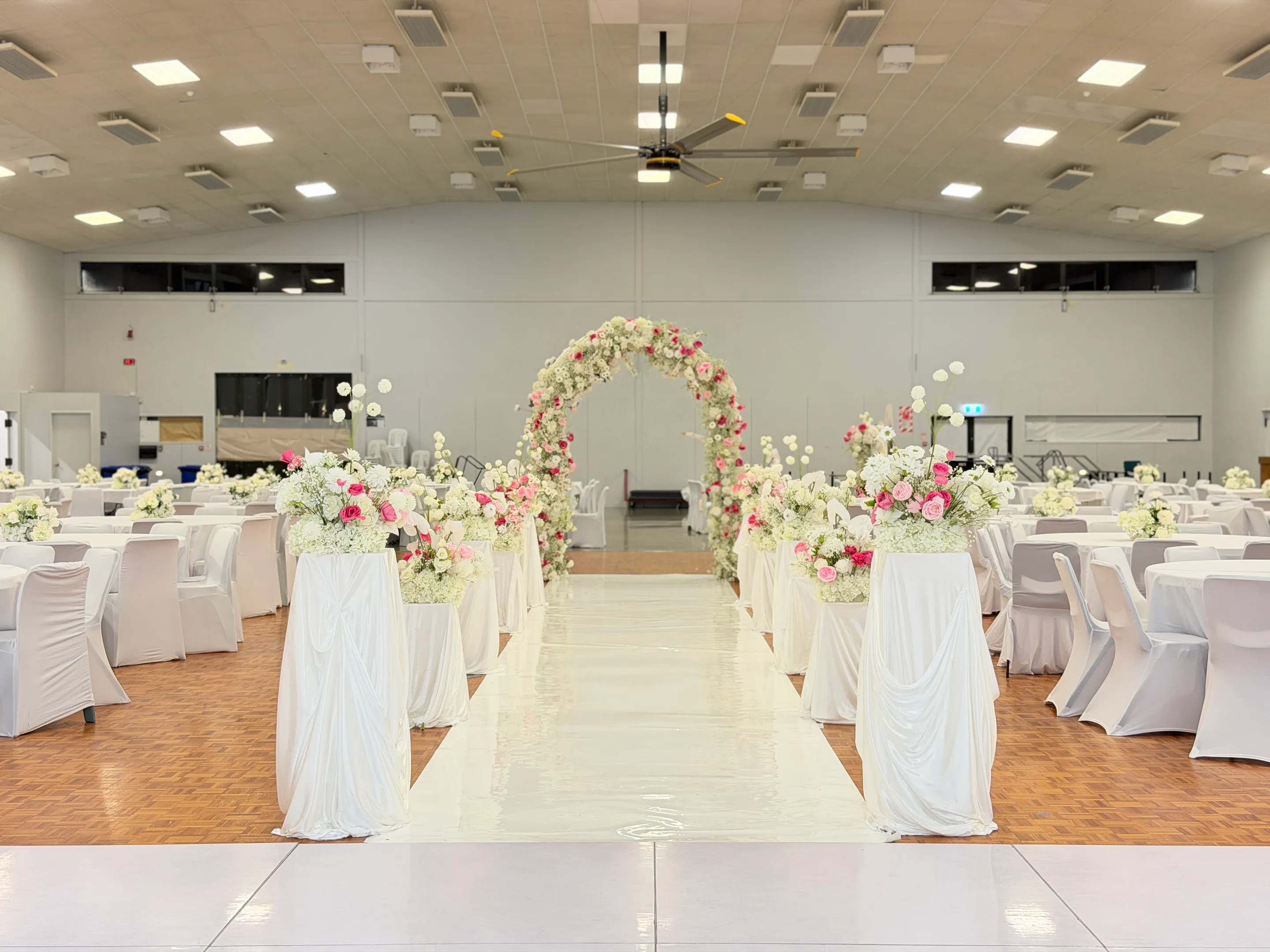 Reception styling by Decoration X: White gloss aisle between draped pedestals of pink and white roses leading to a floral arch, round tables and white-covered chairs on parquet.