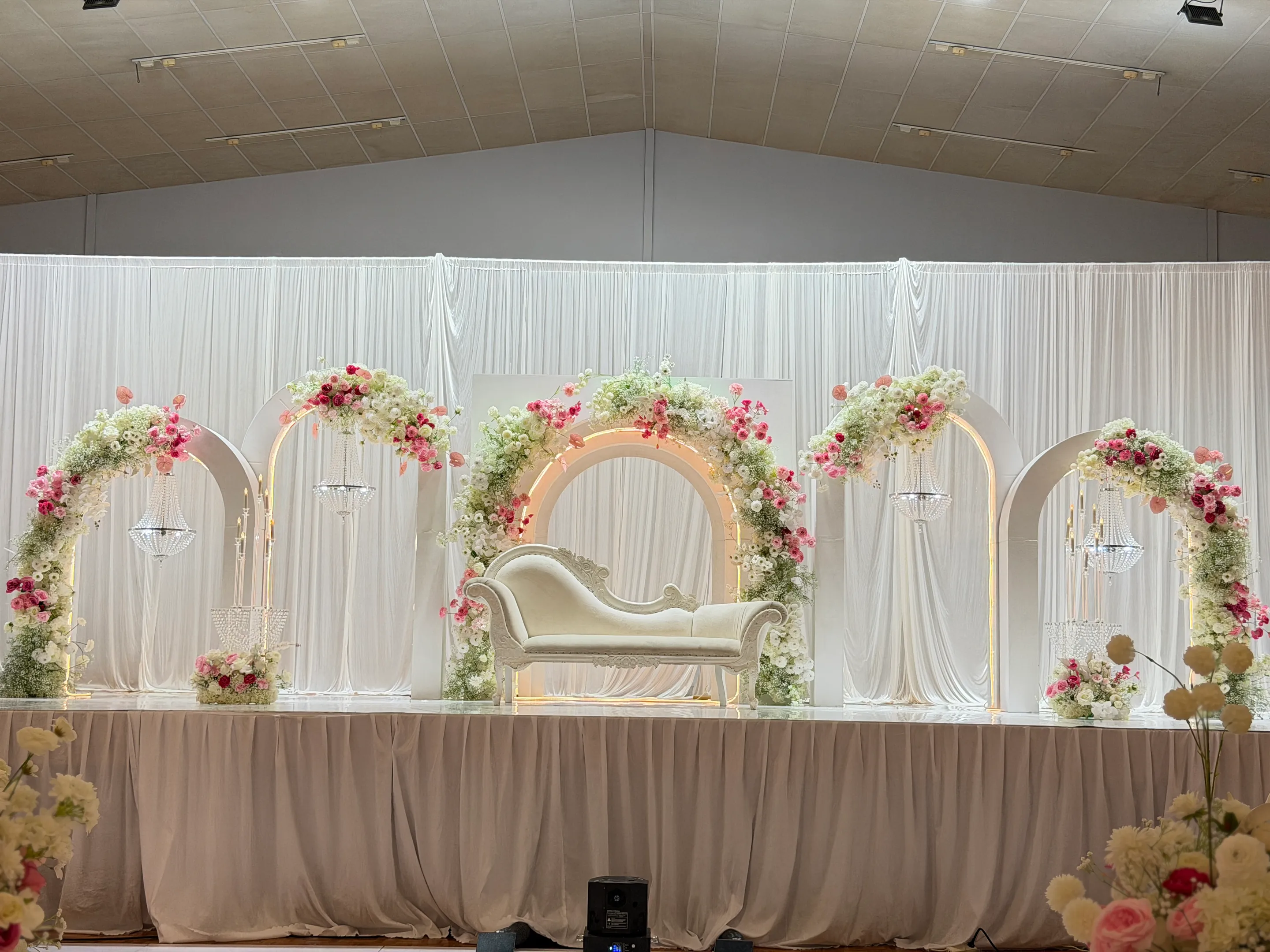Stage styling by Decoration X: Stage of five lit white arches trailing pink and white roses and gypsophila, crystal chandeliers, a carved white chaise at centre, pleated ivory skirting.