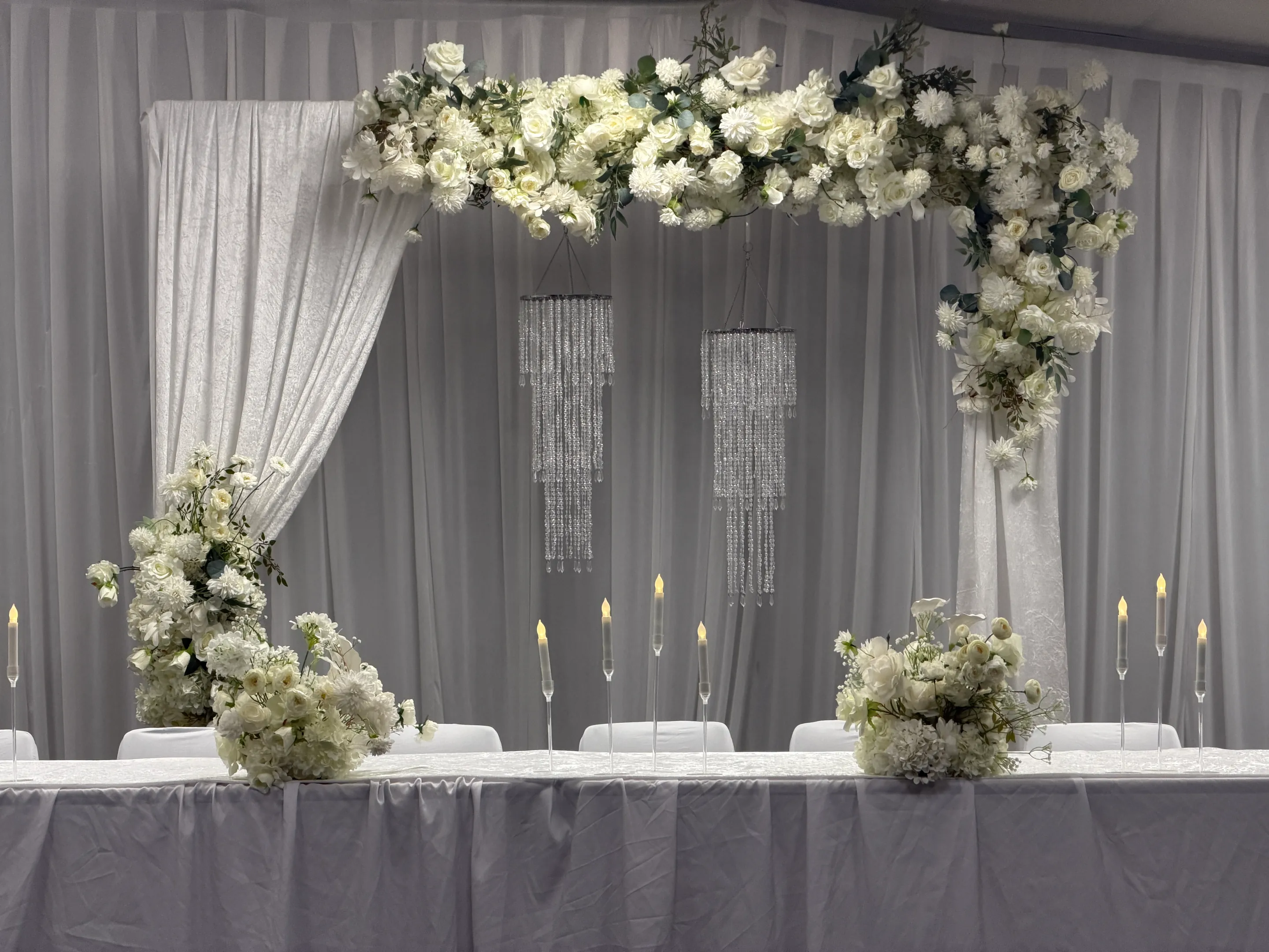 Stage styling by Decoration X: Head table backdrop of white rose and hydrangea swag over ivory drapes, two crystal strand chandeliers, taper candles on glass stems and low white posies.