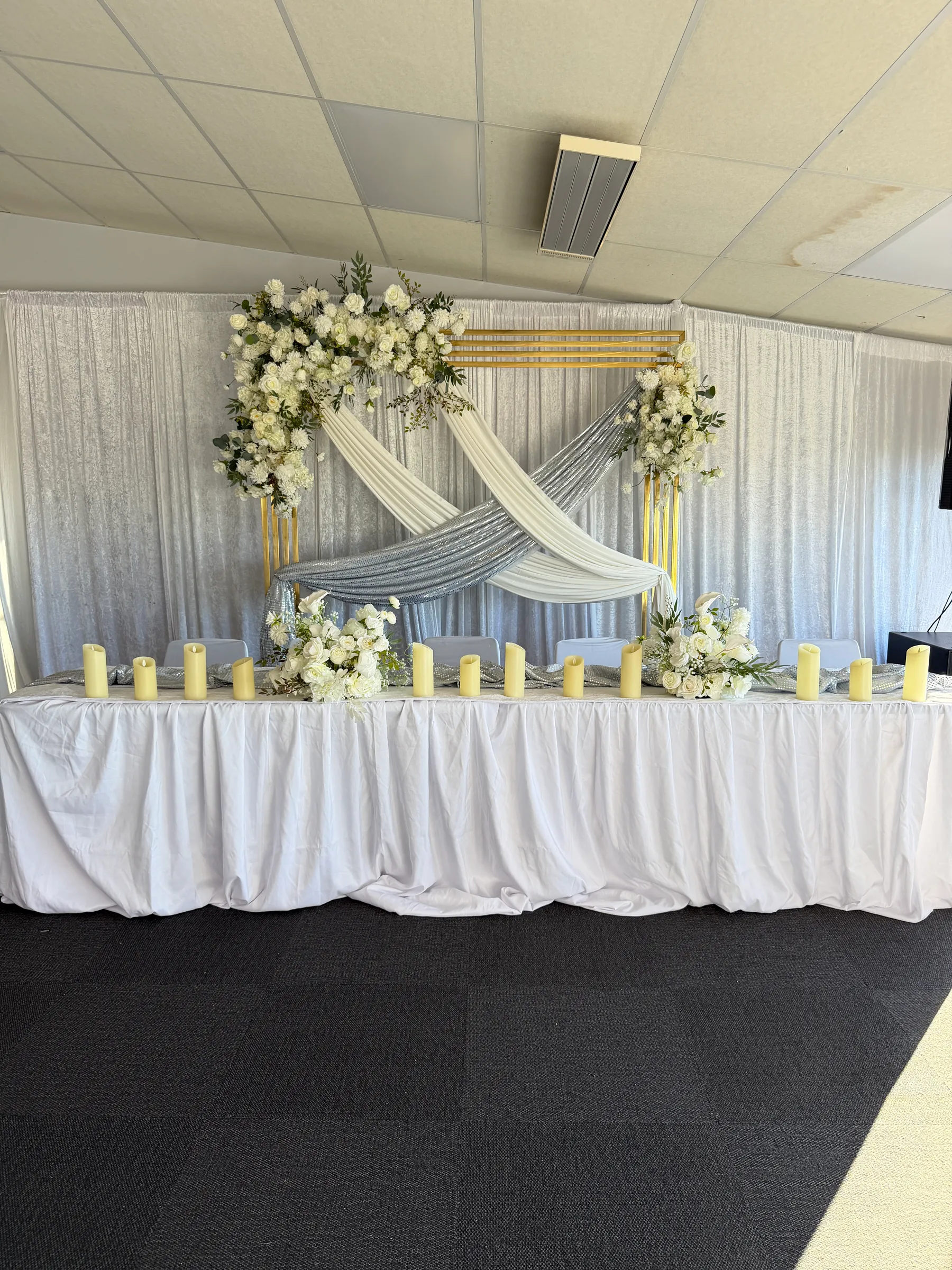 Stage styling by Decoration X: Head table skirted in white with LED pillar candles before a gold frame crossed with ivory and silver drapes, white rose and greenery arrangements.