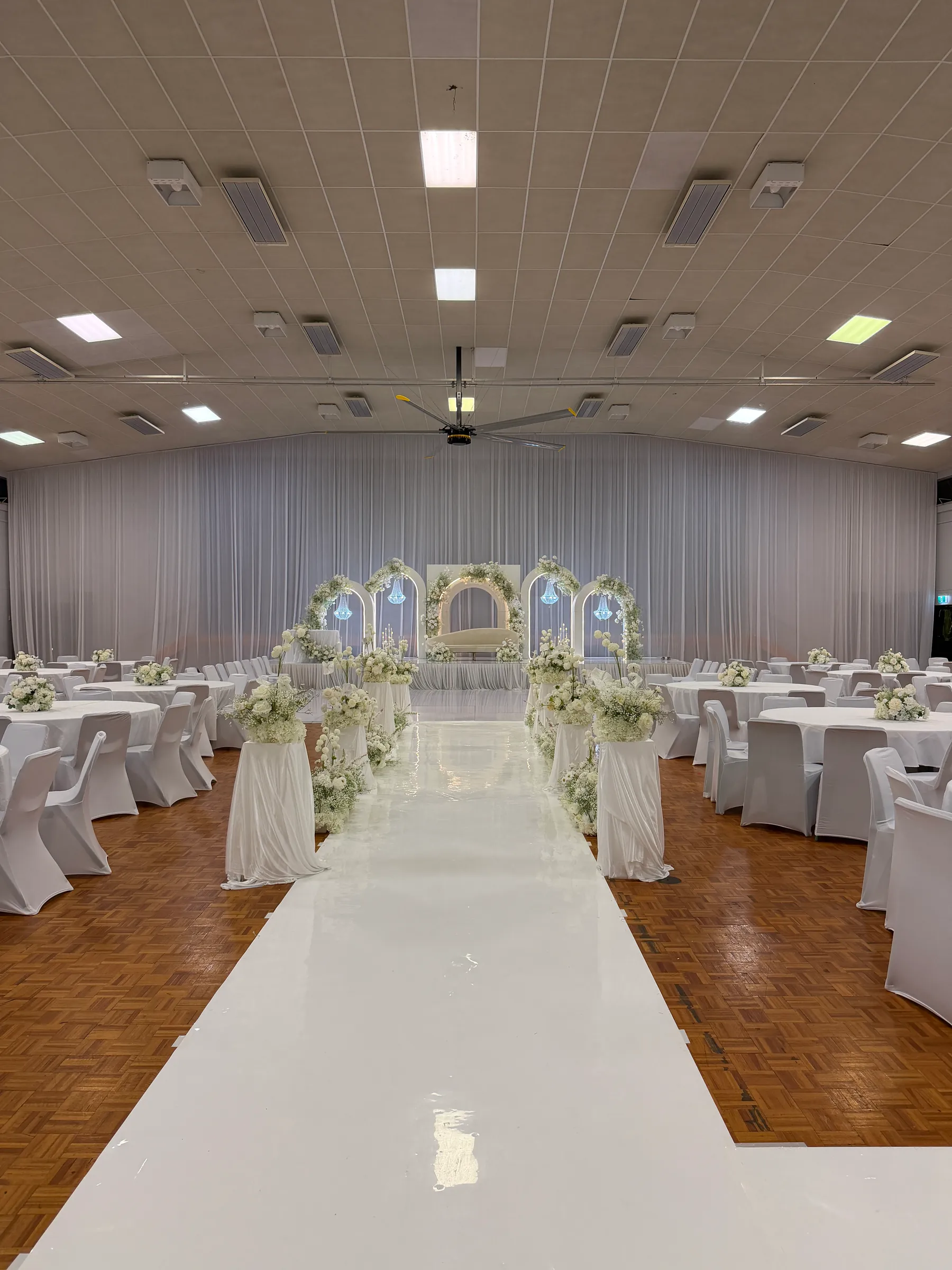 Reception styling by Decoration X: White gloss aisle lined with draped floral pedestals leading to a stage of lit arches, round tables and white-covered chairs filling a hall hung with white drapes.