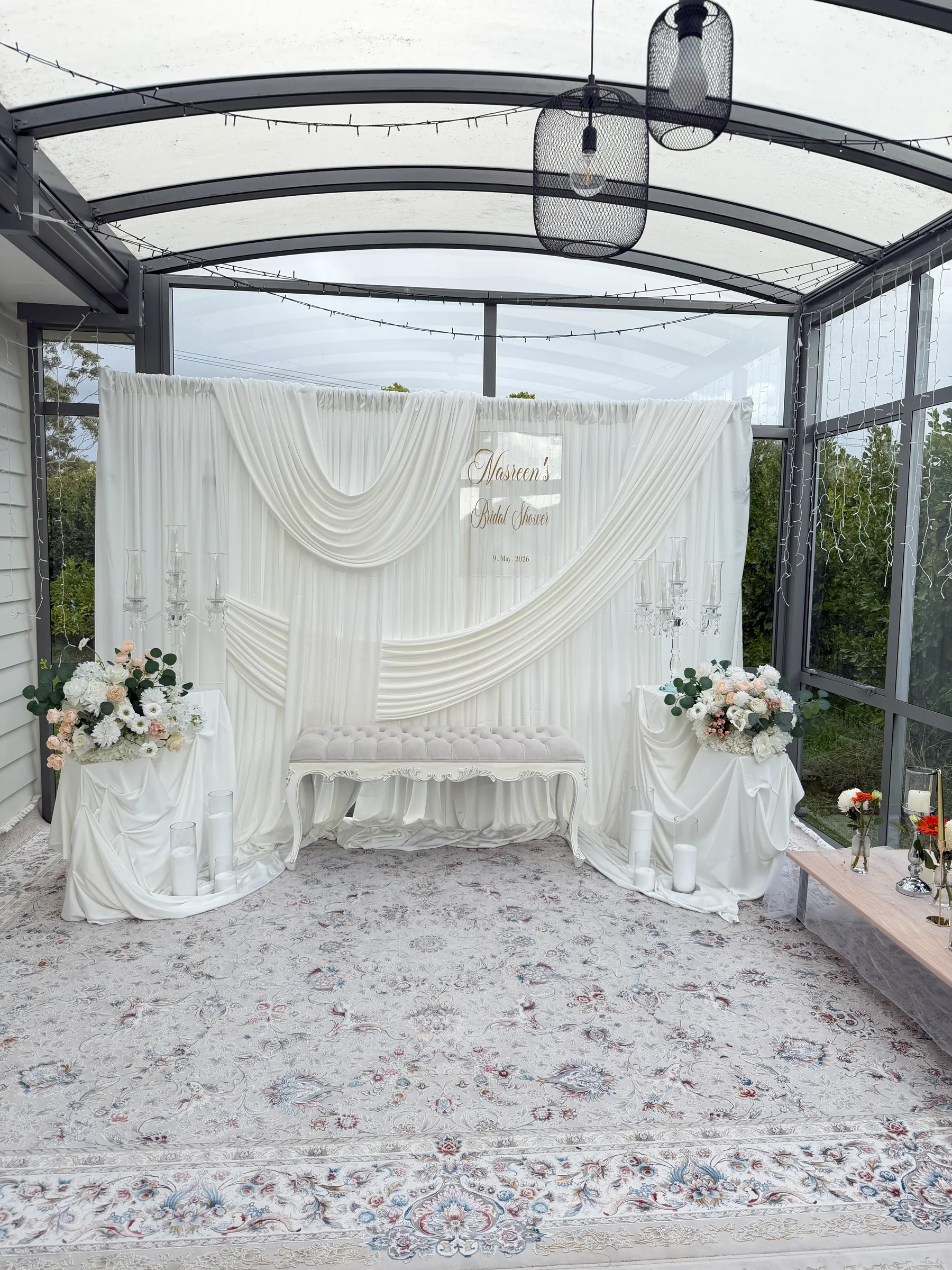 Stage styling by Decoration X: Ivory swagged backdrop with an acrylic name sign, tufted bench, white and blush floral plinths and candelabra on a Persian rug under a glass canopy.
