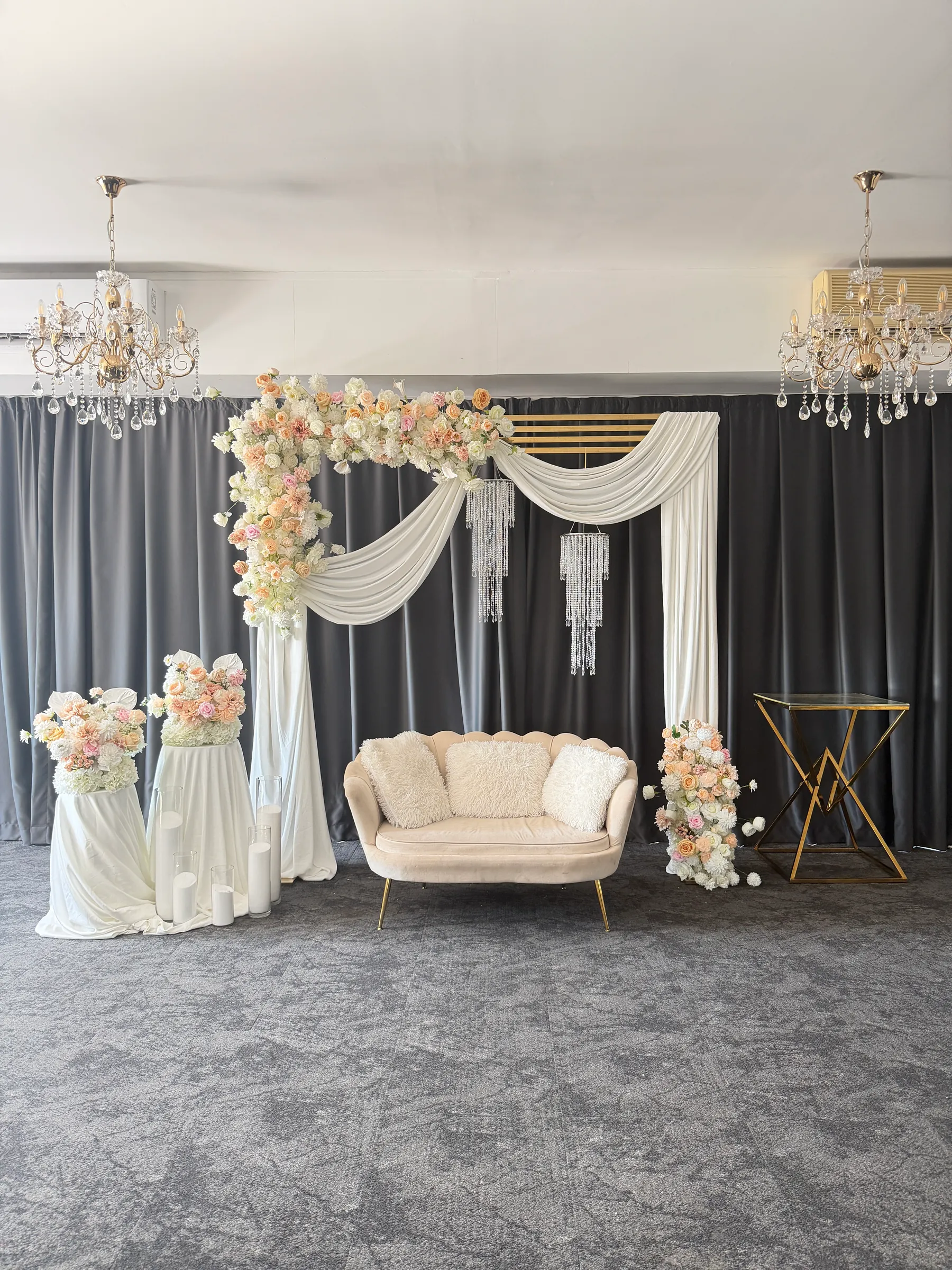 Stage styling by Decoration X: Cream scalloped loveseat beneath a gold arch swagged in ivory chiffon with peach and blush roses, crystal strands, floral plinths and twin chandeliers on grey drapes.