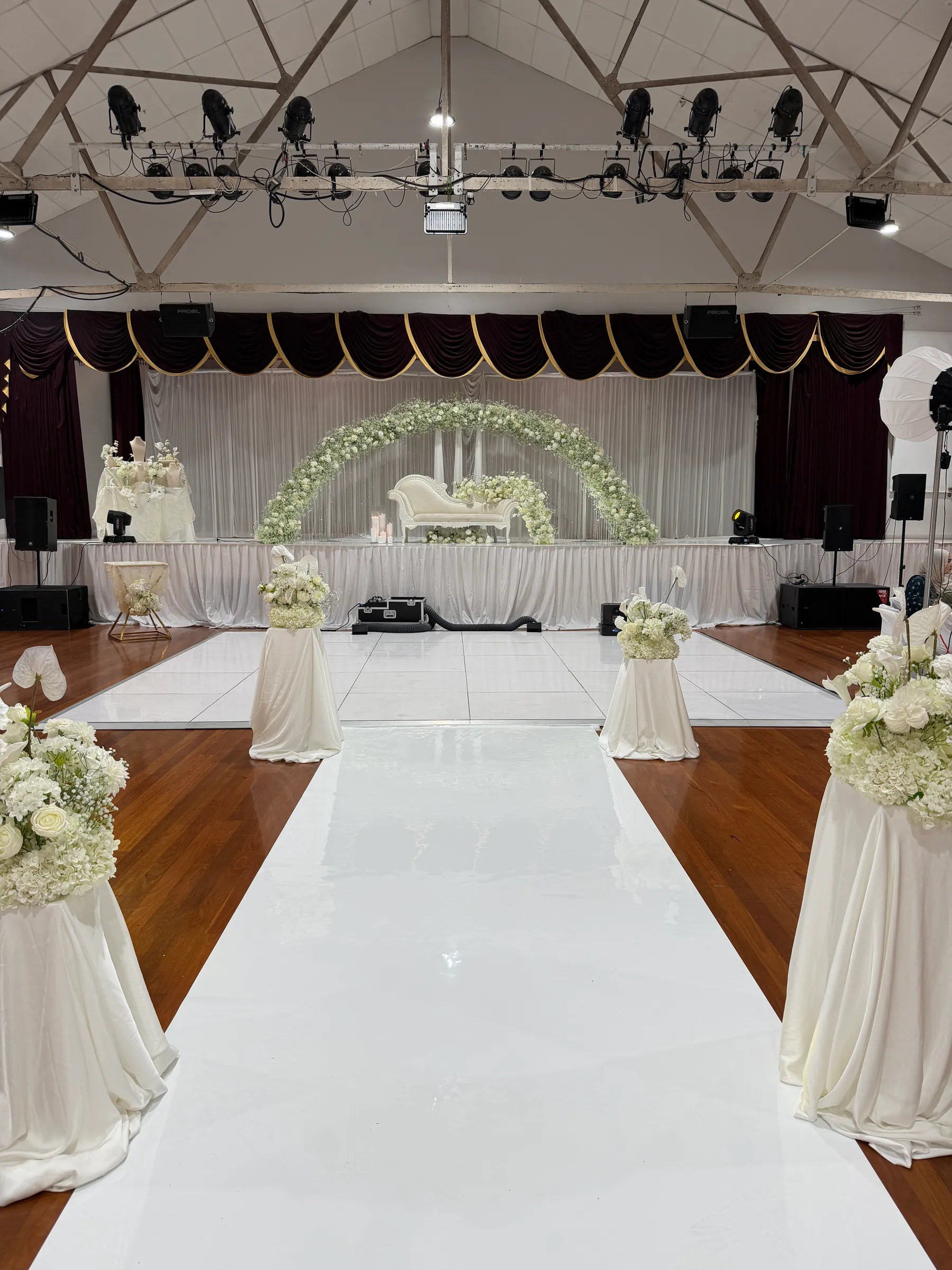 Ceremony styling by Decoration X: Glossy white aisle runner between draped plinths of white flowers, crossing a white dance floor to a gypsophila-arched sofa stage.