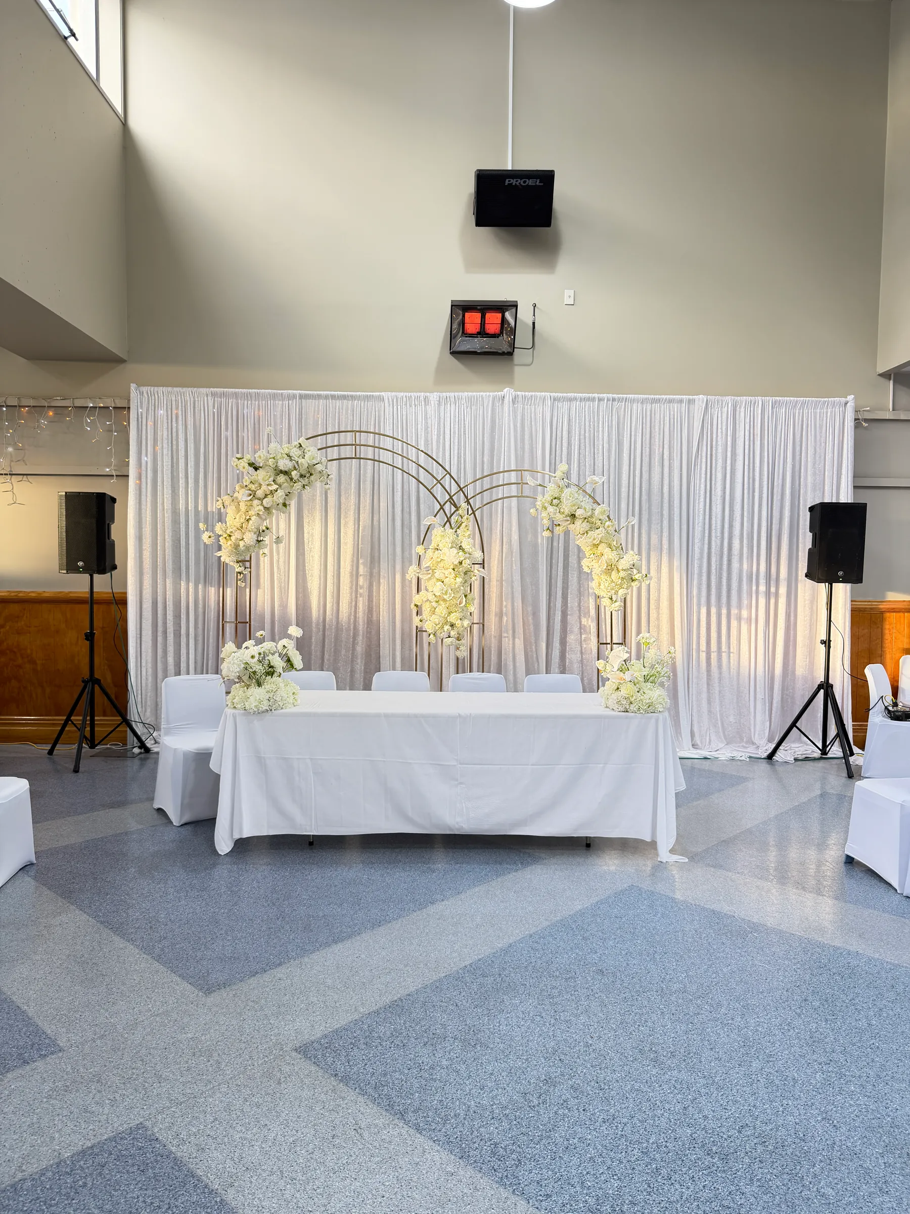 Stage styling by Decoration X: Long white-clothed head table before white drapes and three interlocking gold arches dressed with white flowers, speakers on stands either side.