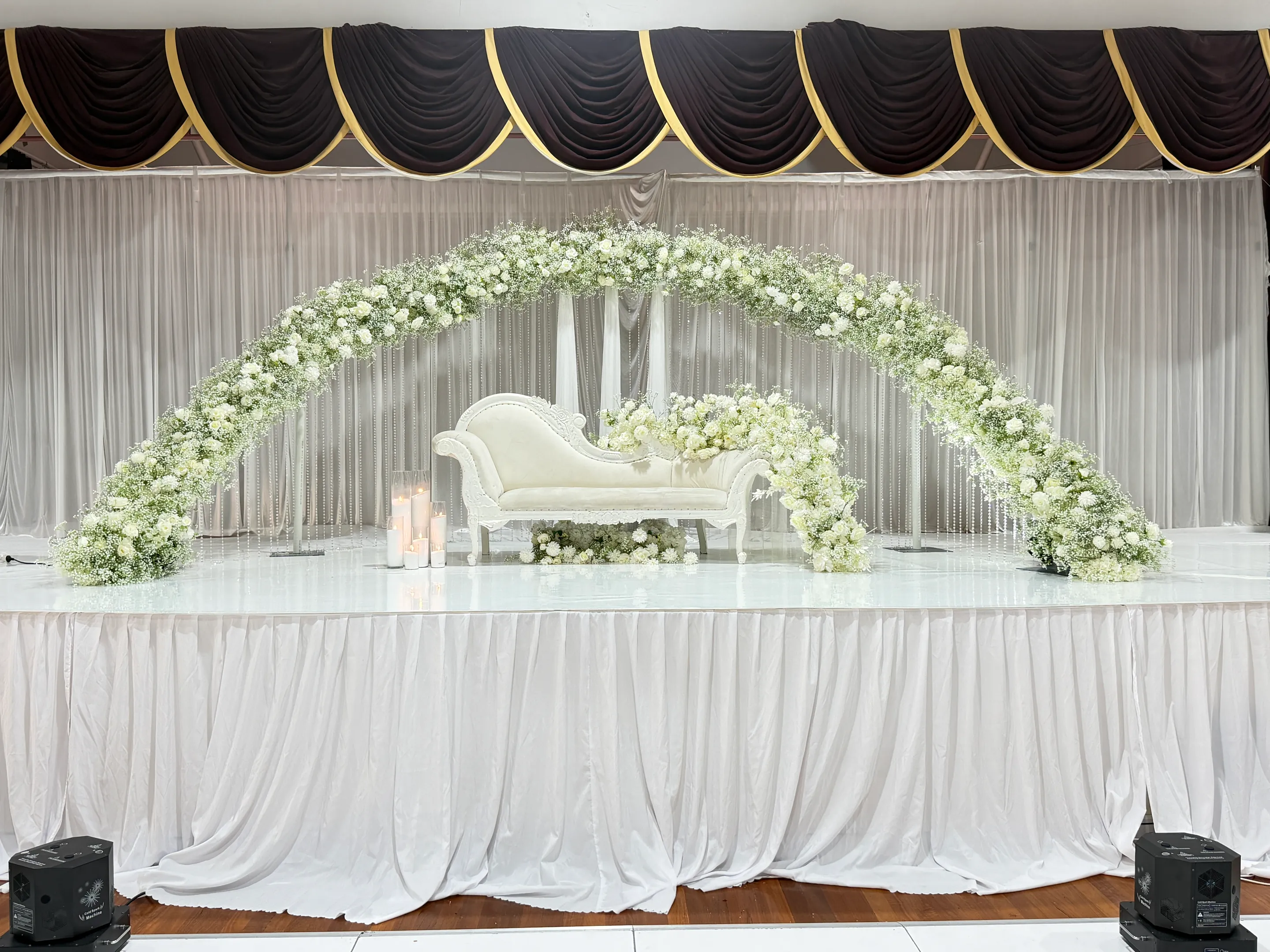 Stage styling by Decoration X: Wide arch of gypsophila and white roses spanning a white-skirted stage, an ivory chaise and pillar candles beneath, black and gold pelmet above.