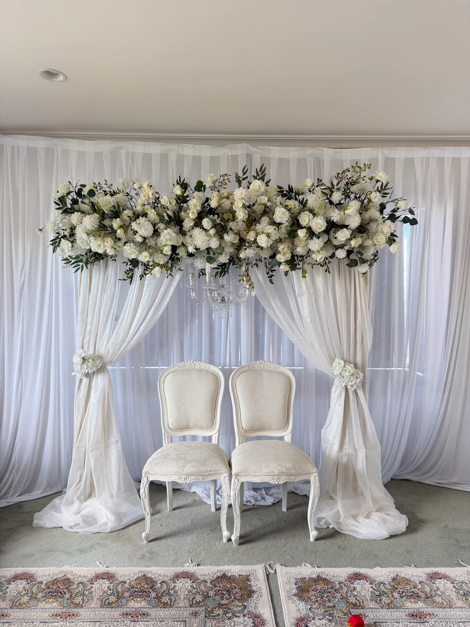 Stage styling by Decoration X: Two ivory upholstered chairs beneath a long swag of white roses and greenery, sheer drapes tied back with flowers, Persian rugs in front.