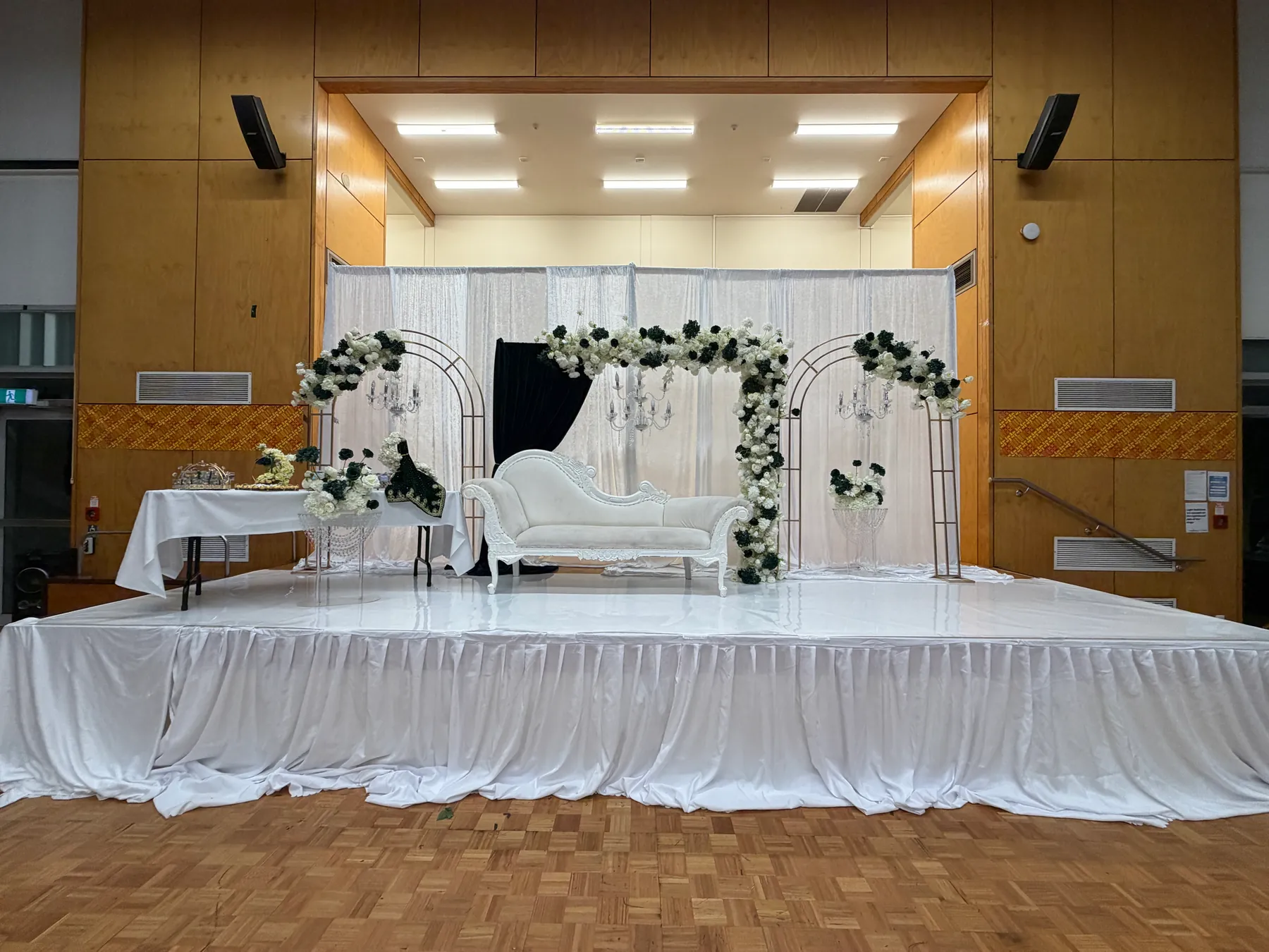 Stage styling by Decoration X: Ivory chaise on a white-skirted stage framed by gold arches dressed in white and deep-green flowers, crystal candelabra and a draped side table.