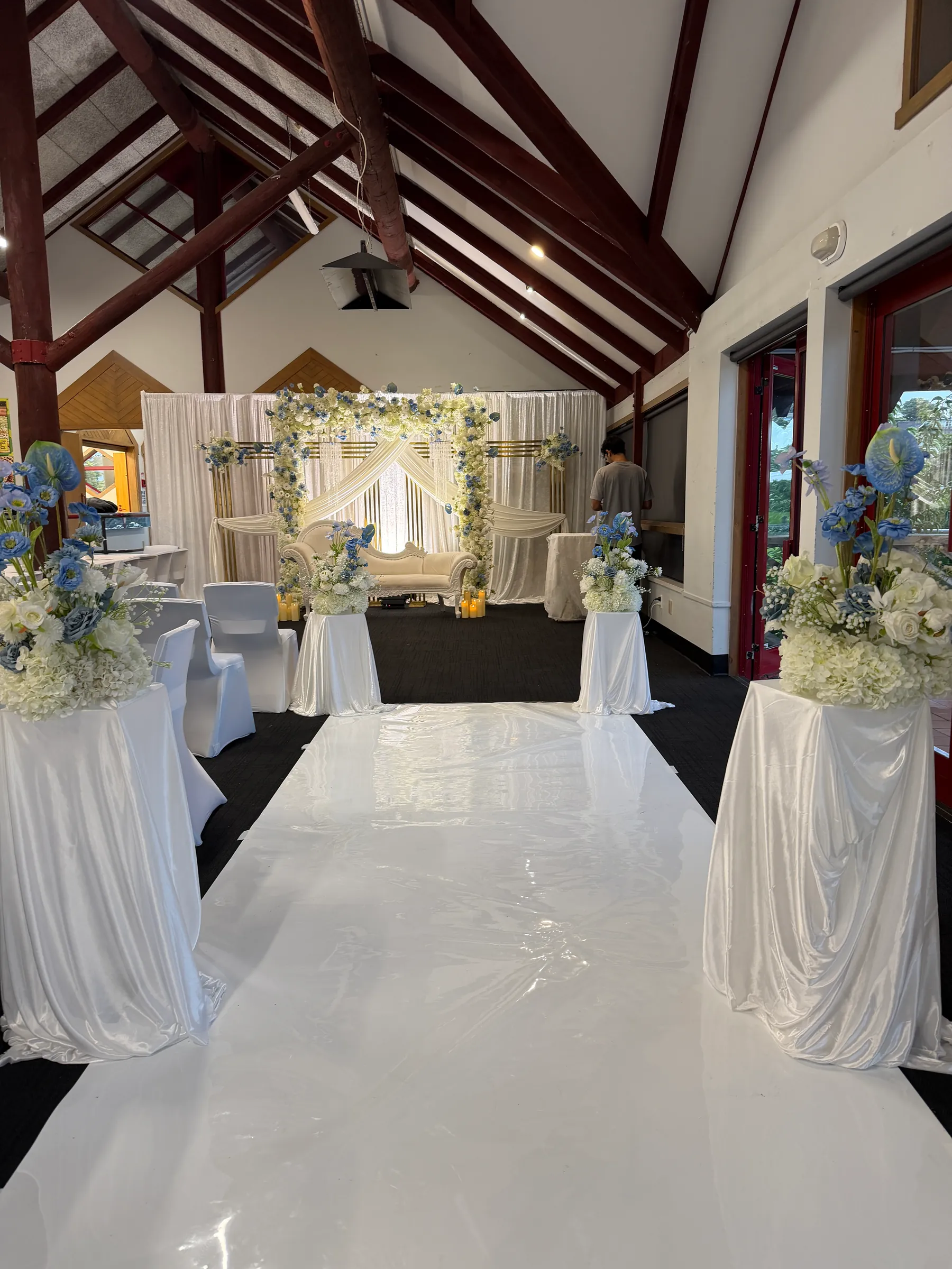 Ceremony styling by Decoration X: Glossy white aisle runner between draped plinths topped with blue and white hydrangea arrangements, leading to a floral-arched sofa stage.