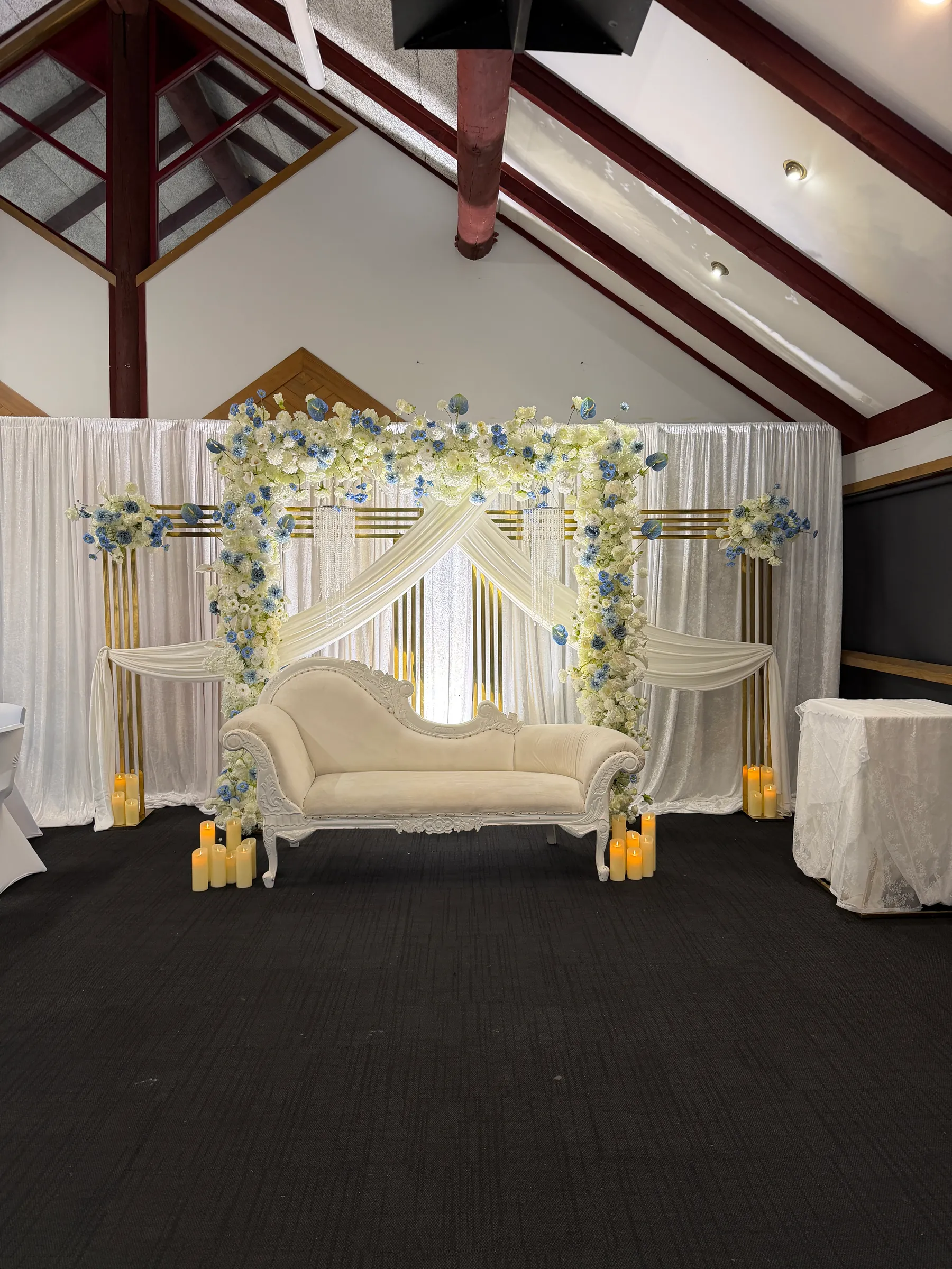 Stage styling by Decoration X: Ivory chaise under a gold-framed arch thick with white and blue flowers, swagged white drapes, gold slats and pillar candles at its feet.