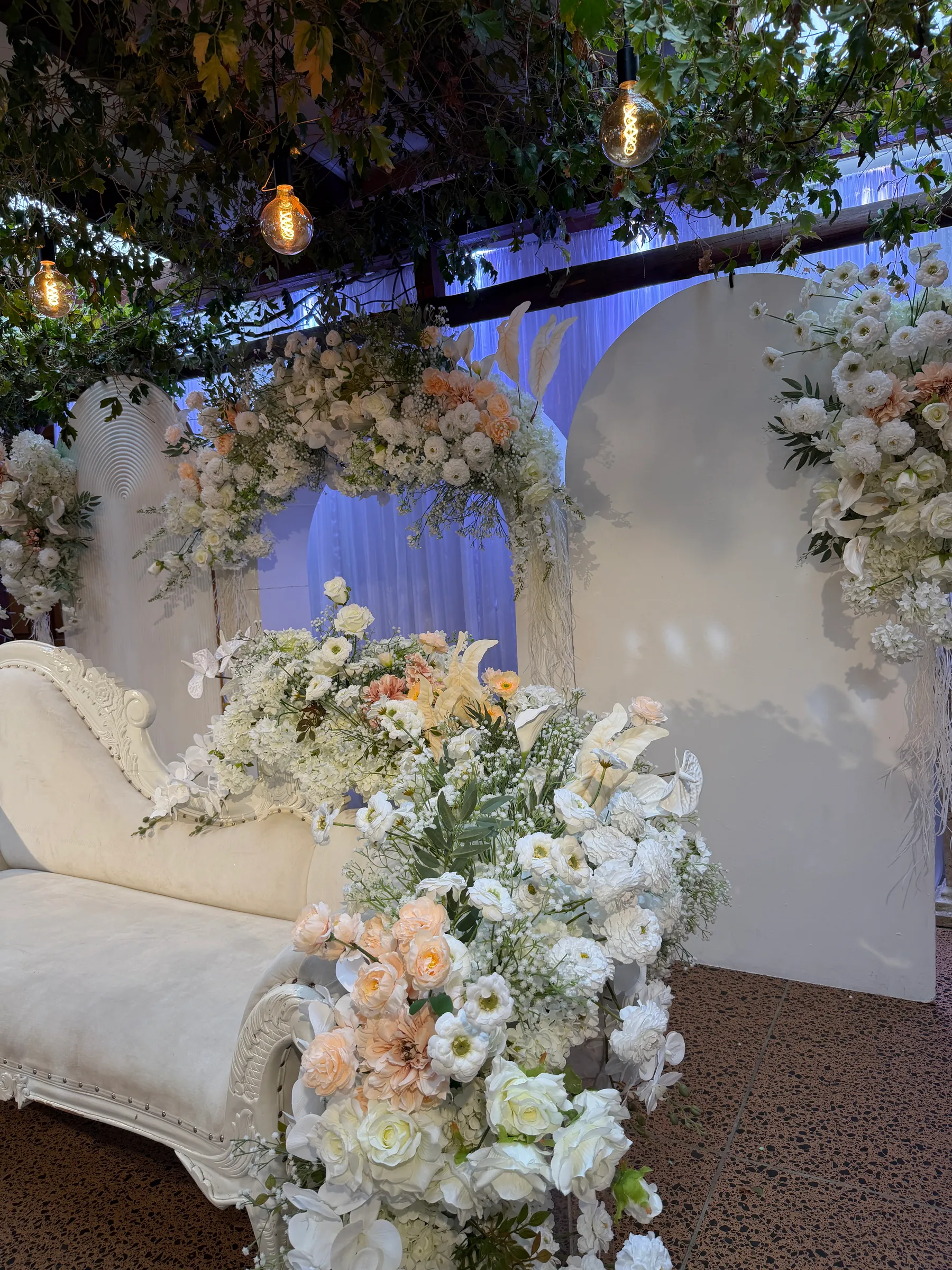 Stage styling by Decoration X: Close on white roses, peach dahlias, anthuriums and gypsophila spilling over an ivory chaise arm, white arched panels and Edison bulbs behind.