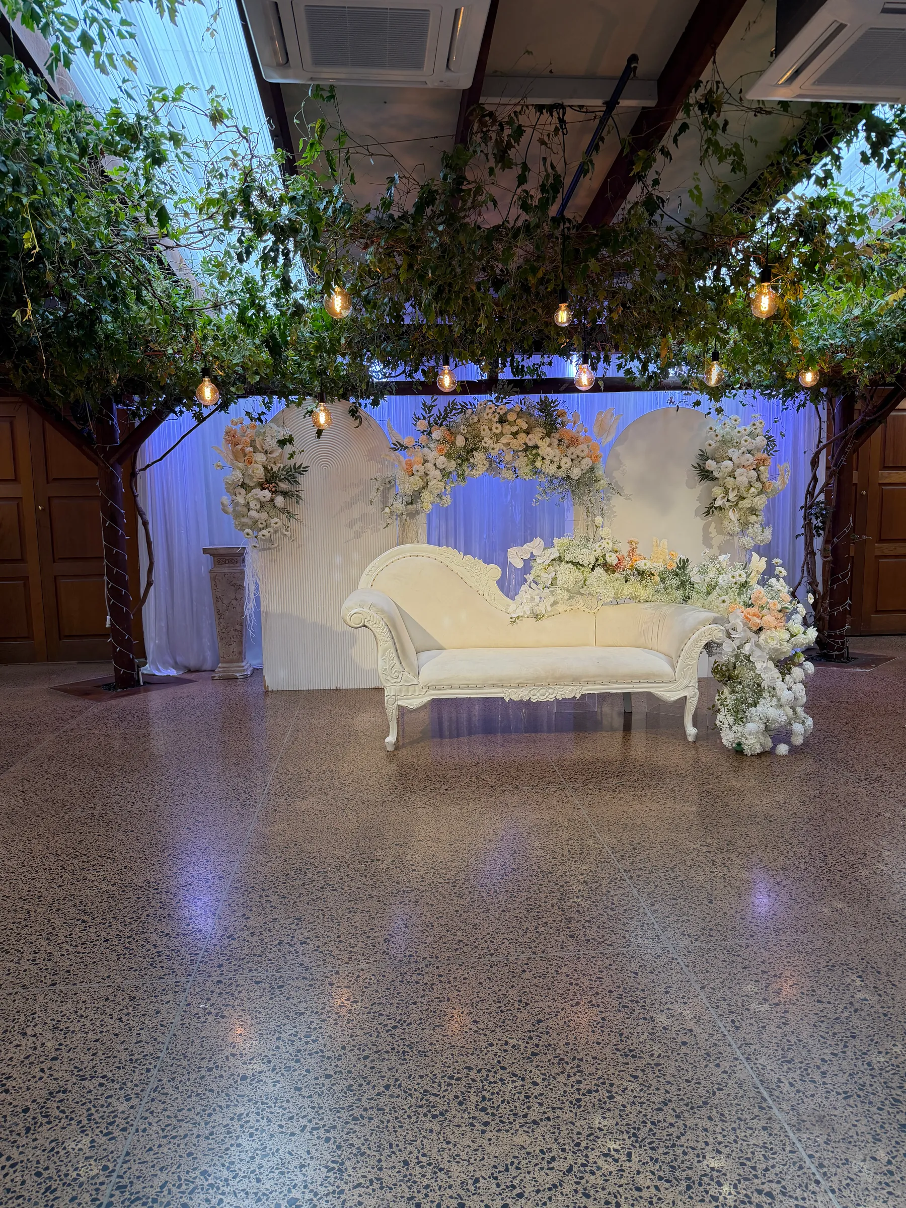 Stage styling by Decoration X: Ivory carved chaise beneath an arch of white and peach roses between two white arched panels, a vine canopy with festoon bulbs overhead.