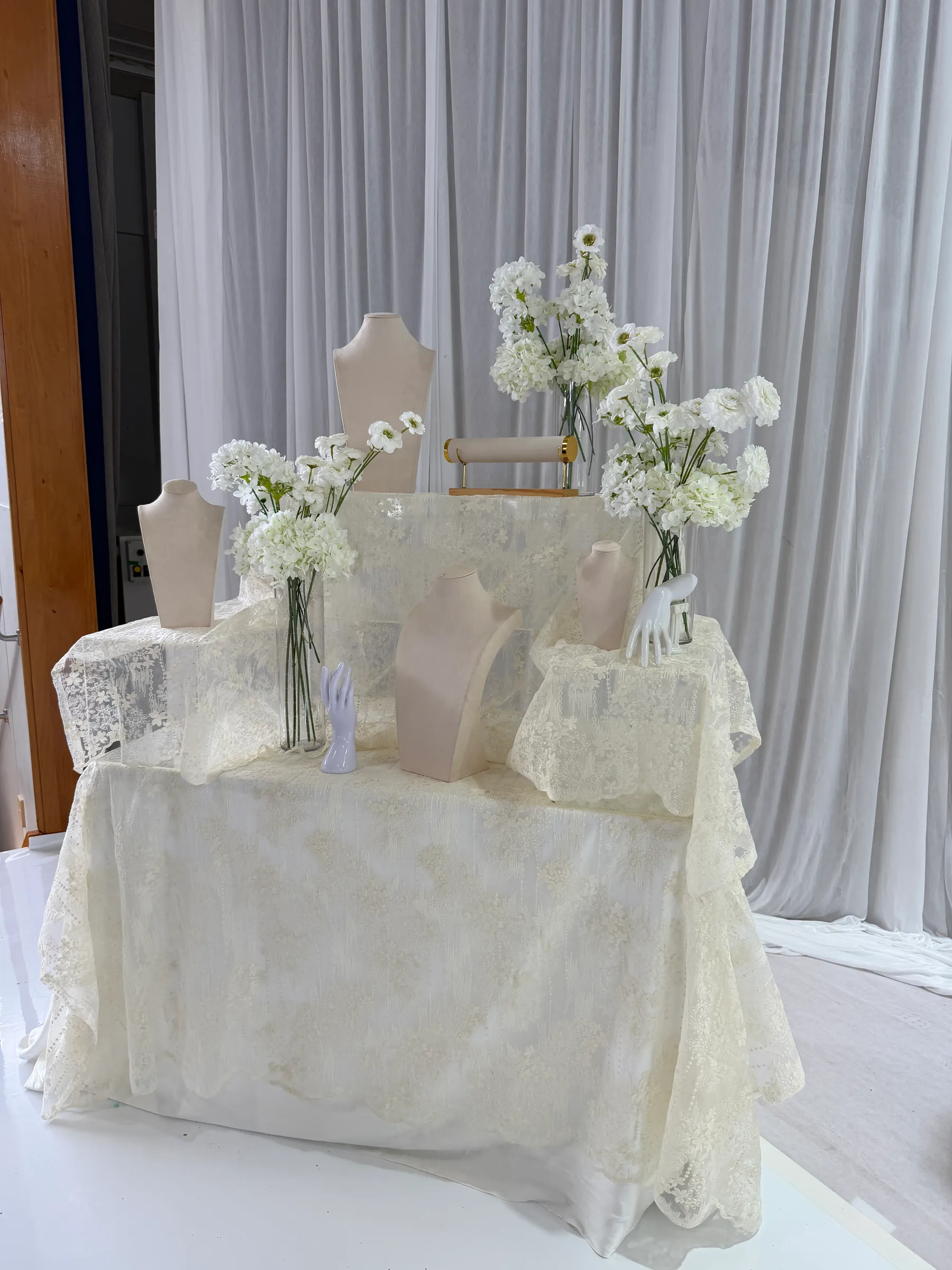 Tables styling by Decoration X: Tiered display table in ivory lace with velvet jewellery busts, hand stands, a bracelet bar and vases of white blooms, against white drapes.