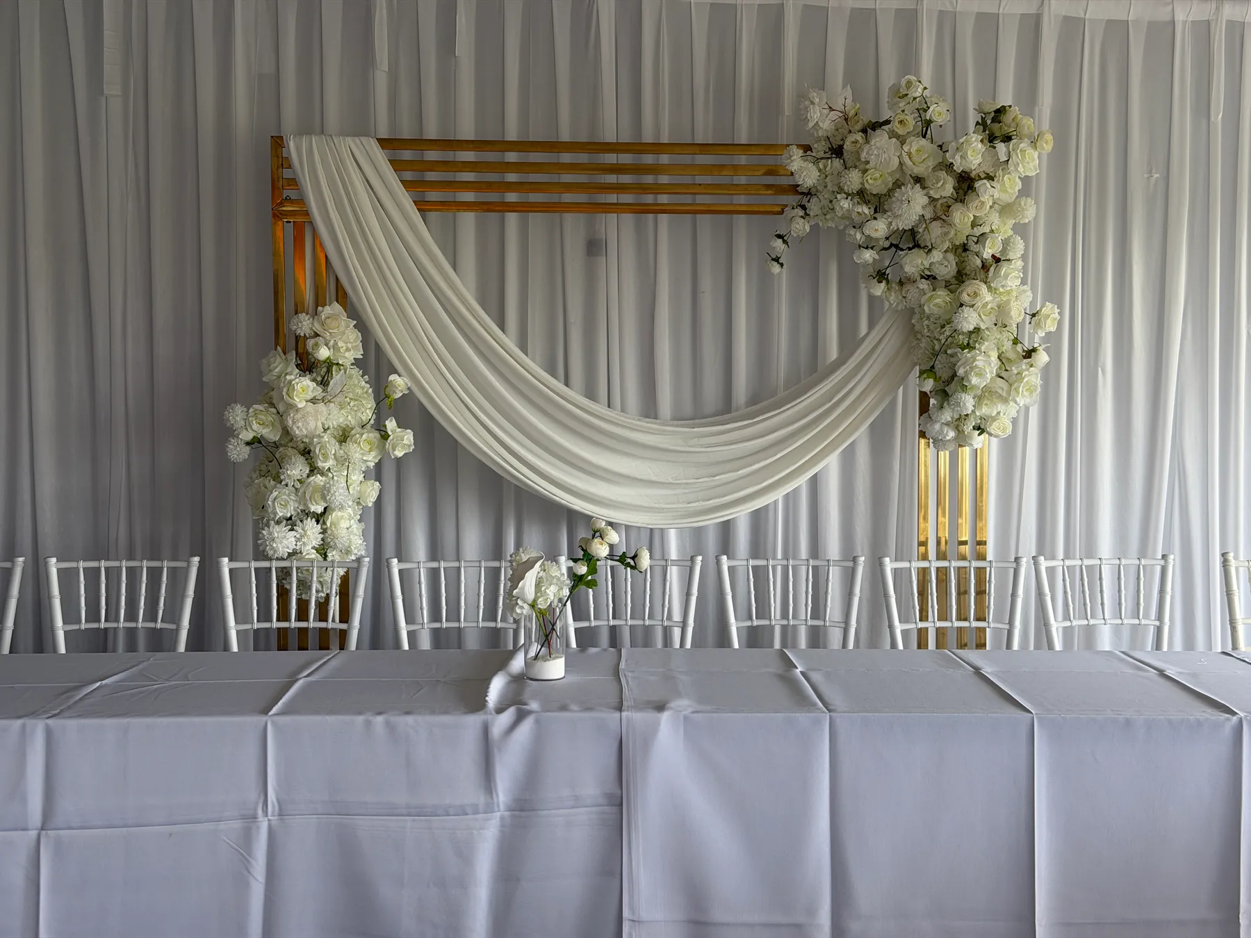 Stage styling by Decoration X: Gold slatted frame with an ivory chiffon swag and white rose and chrysanthemum clusters behind a long head table and a row of white chiavari chairs.