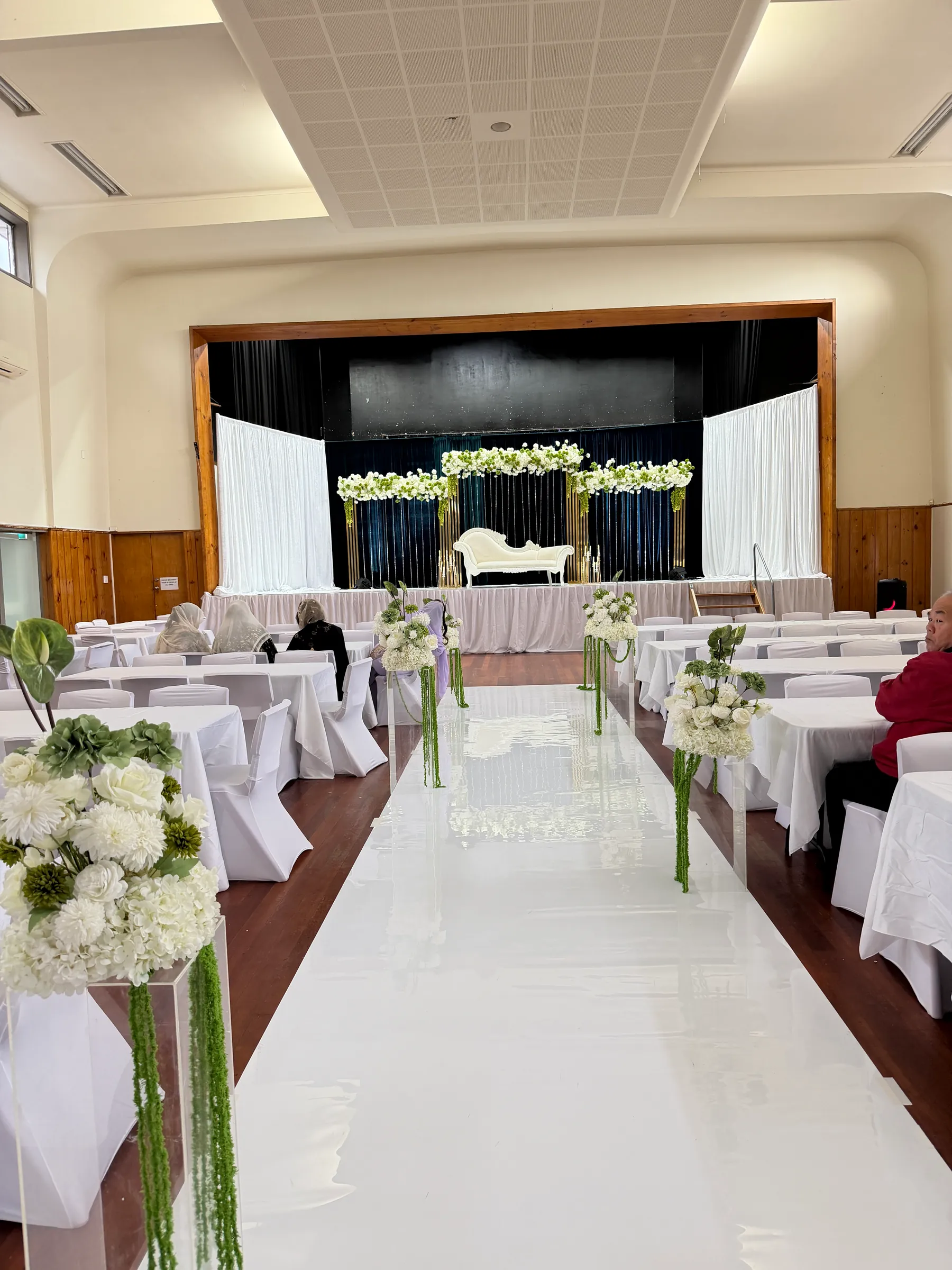 Ceremony styling by Decoration X: White aisle runner lined with acrylic plinths of white hydrangea and trailing green amaranthus, leading past white-clothed tables to a teal and gold stage.