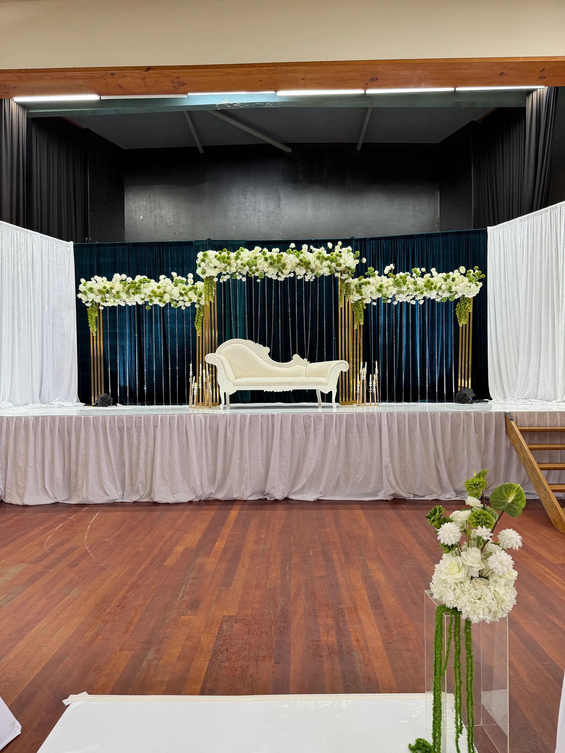 Stage styling by Decoration X: Teal velvet stage backdrop hung with crystal strands, gold frames under white and green floral garlands, a white chaise, a white hydrangea plinth in front.