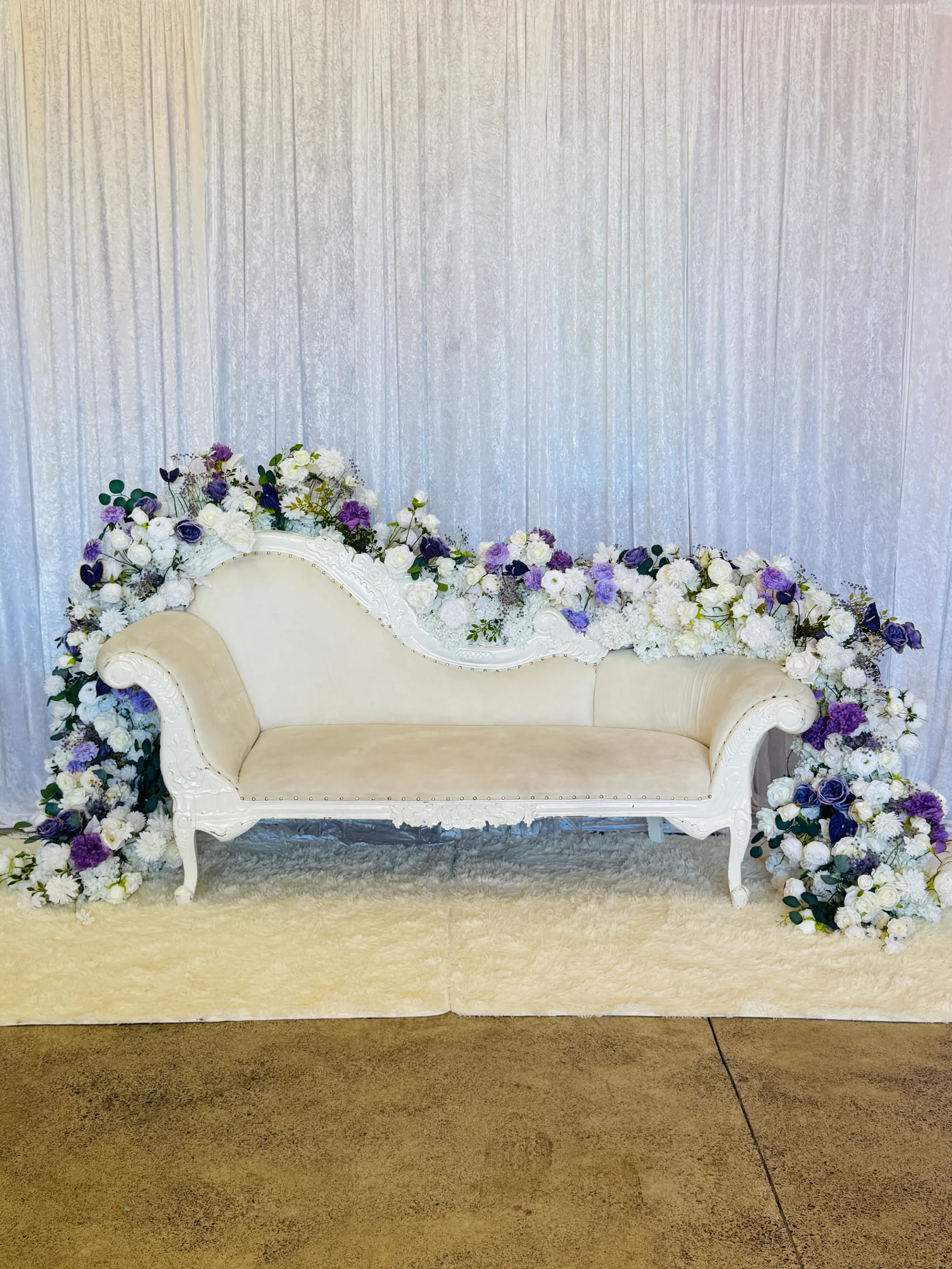 Stage styling by Decoration X: White carved chaise draped end to end with white roses and purple and violet blooms, on a shaggy rug before a crushed white velvet backdrop.