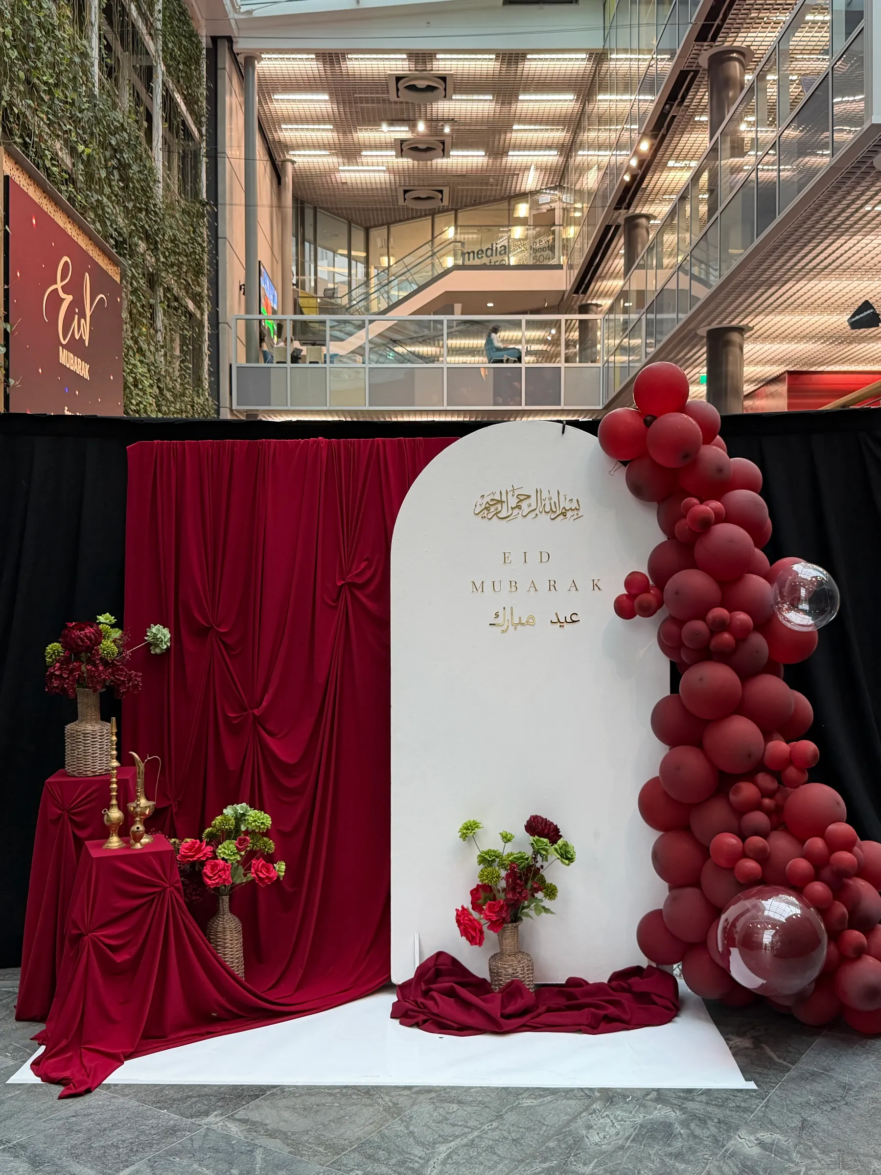 Cultural styling by Decoration X: White arch board lettered Eid Mubarak in gold, a burgundy balloon garland with clear bubble balloons, red velvet drapes and plinths with brass ewers and roses.