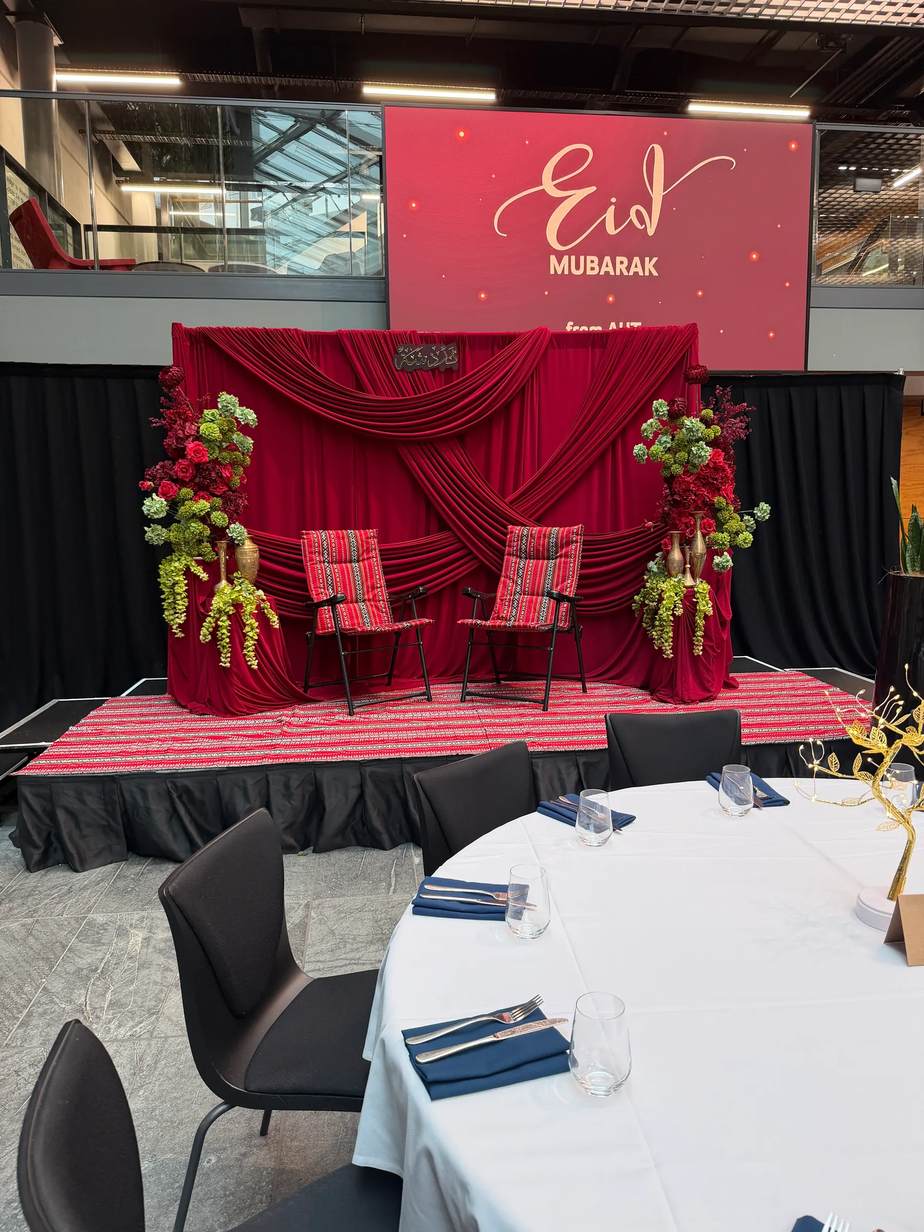 Cultural styling by Decoration X: Red stage backdrop of crossed swags, two red woven-cushion chairs between plinths of red roses, green hydrangea and trailing amaranthus with brass vases.
