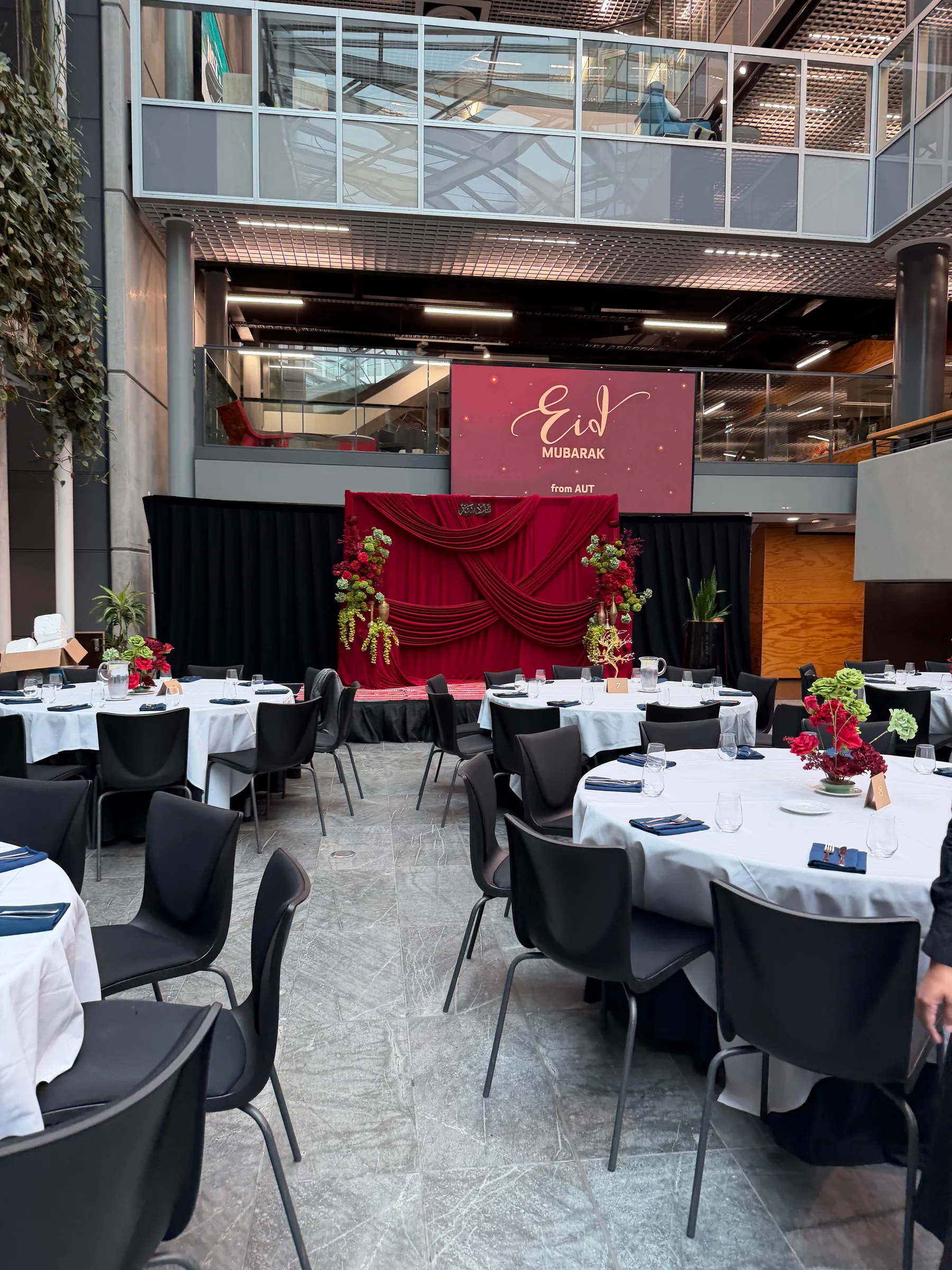 Cultural styling by Decoration X: Atrium dinner of round tables in white linen with navy napkins and red rose centrepieces, facing a red-draped stage beneath an Eid Mubarak screen.