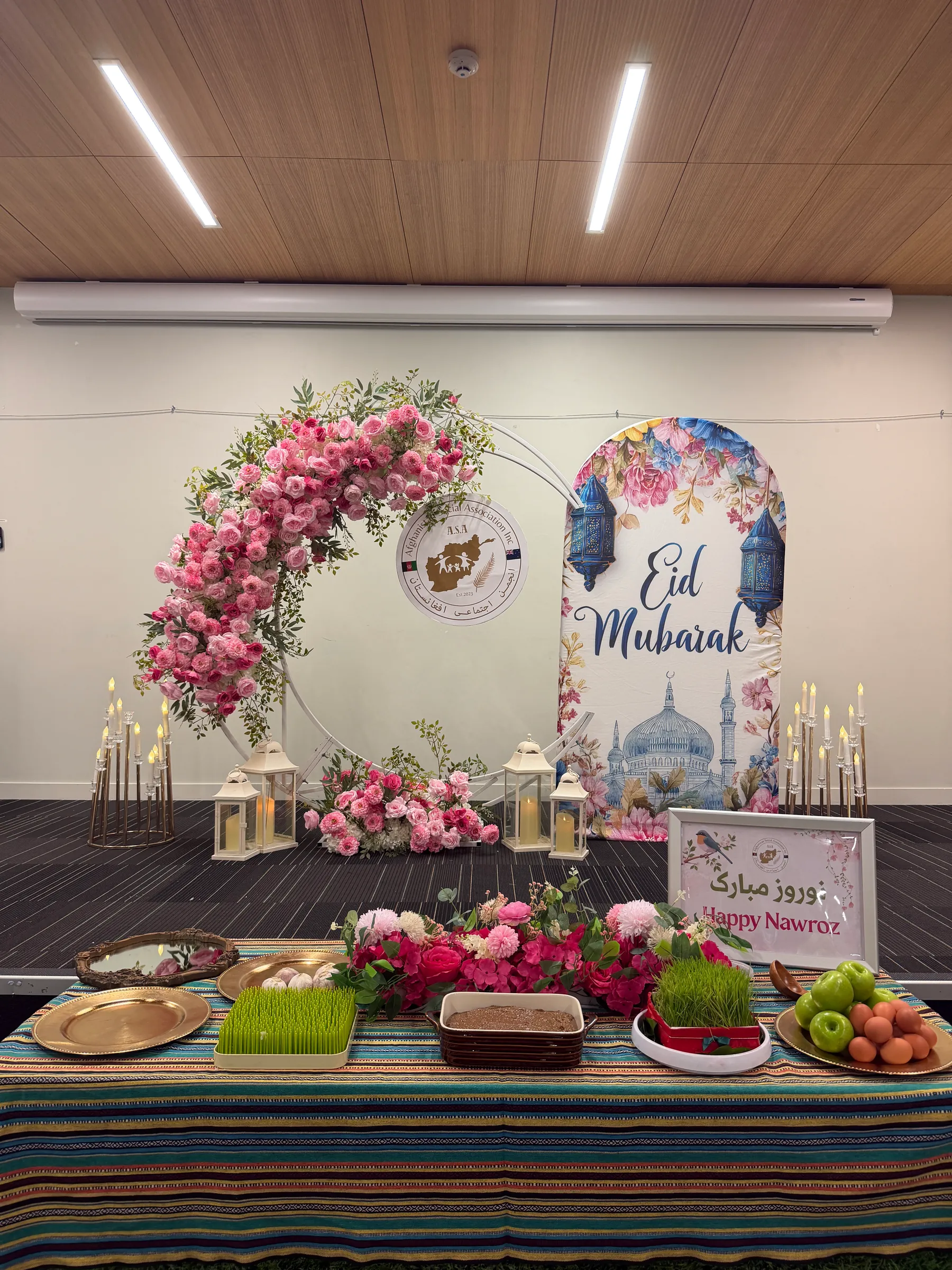 Cultural styling by Decoration X: Pink rose moon arch, Eid Mubarak arch panel and gold candelabra behind a striped-cloth table of wheatgrass, a pudding dish, fruit and a Happy Nawroz sign.