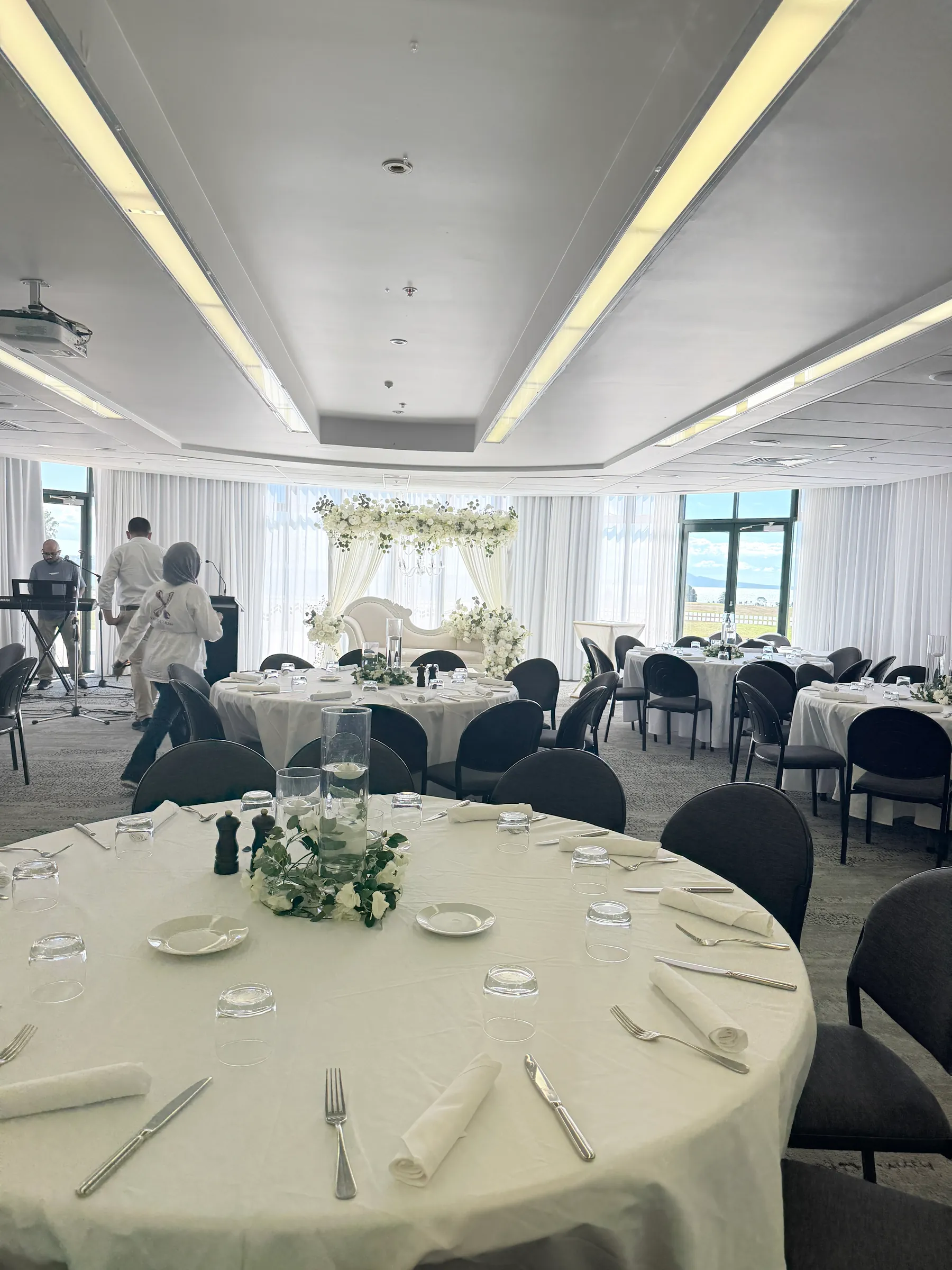Reception styling by Decoration X: Round tables in white linen with floating-candle cylinders and white floral rings, the white chaise stage at the far end before sea-view windows.