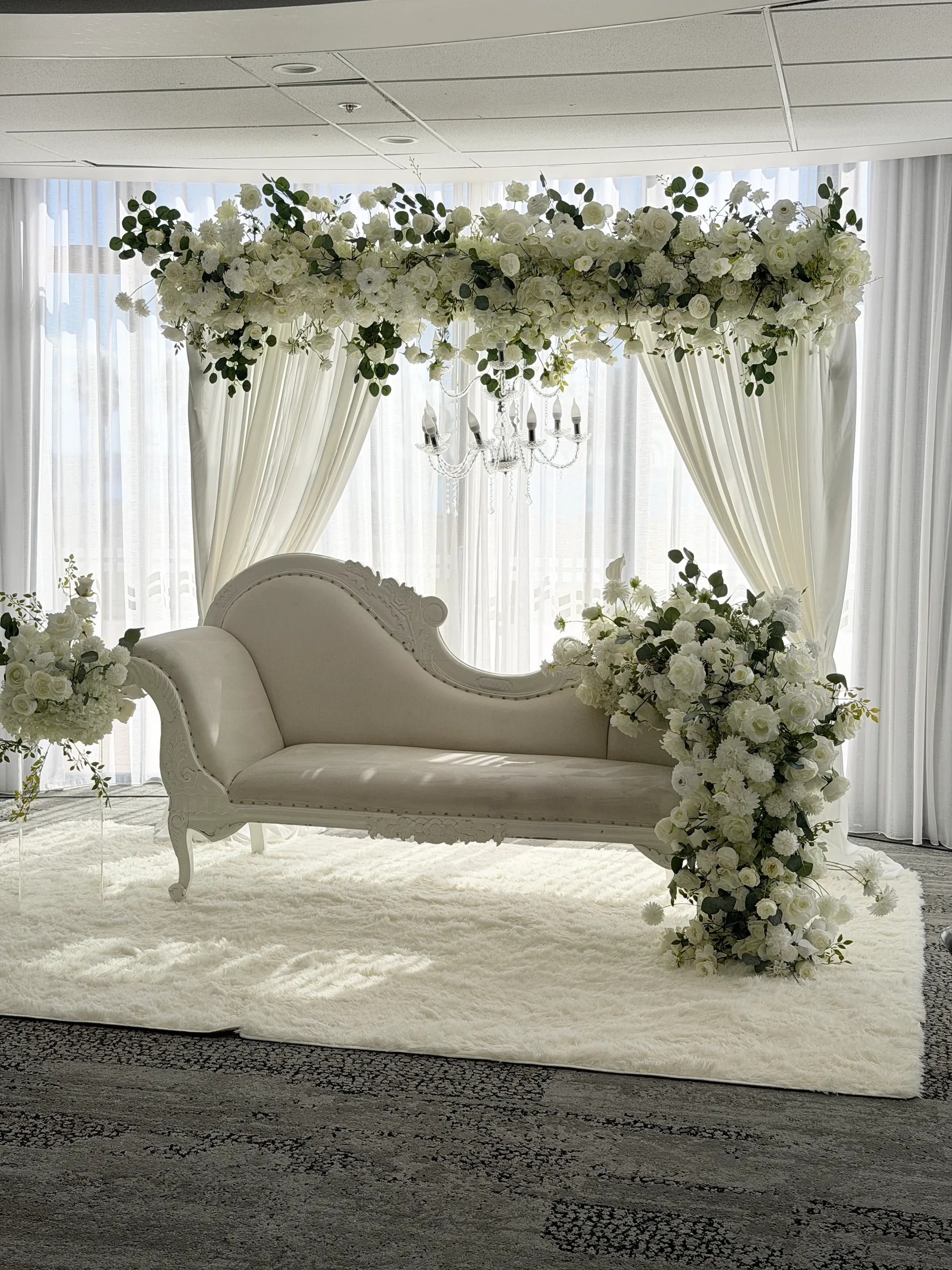 Stage styling by Decoration X: White carved chaise on a shaggy rug beneath a white rose and eucalyptus canopy with a crystal chandelier, sheer drapes and a floral cascade at its arm.
