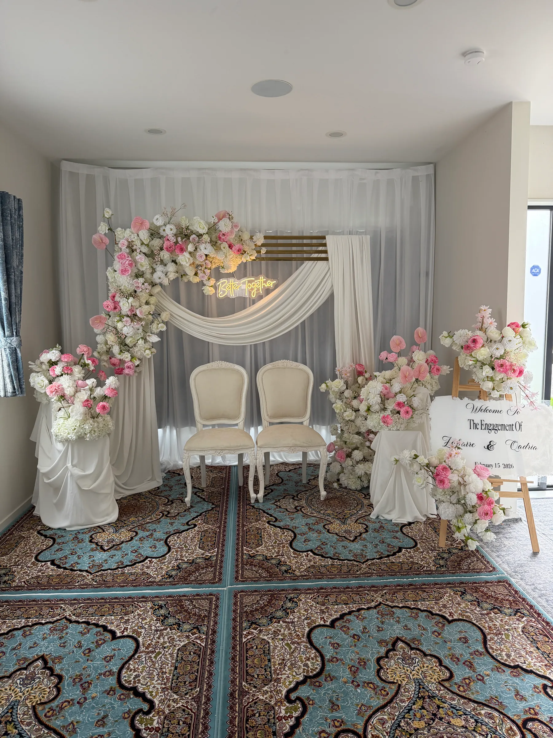 Engagement styling by Decoration X: Pink and white rose arch with a neon script sign, two white French chairs and draped plinths of roses on Persian rugs, a welcome sign on an easel.