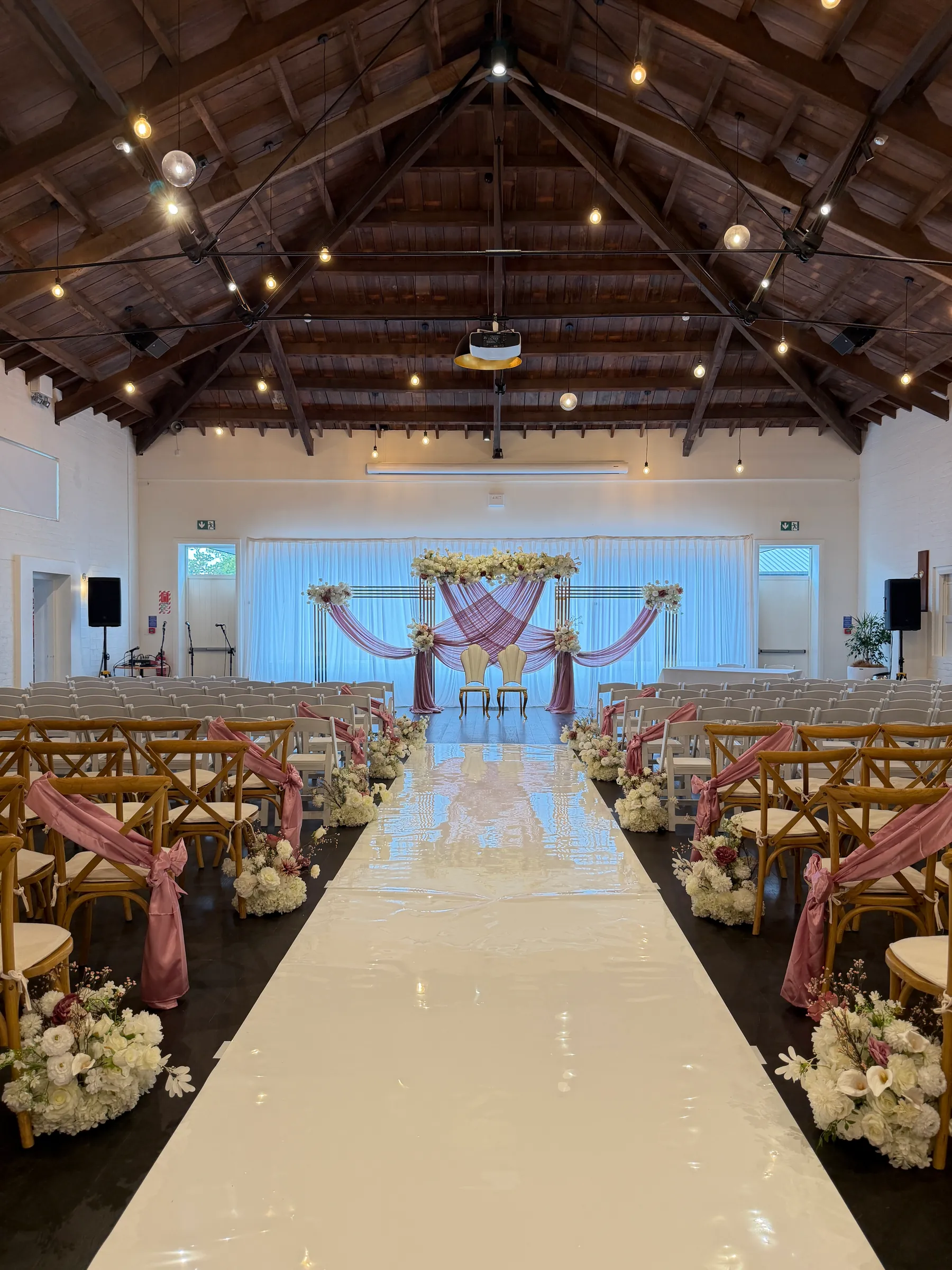 Ceremony styling by Decoration X: White aisle runner between cross-back chairs tied with dusty pink sashes and white floral clusters, leading to a pink-draped gold frame arch under a timber truss ceiling.