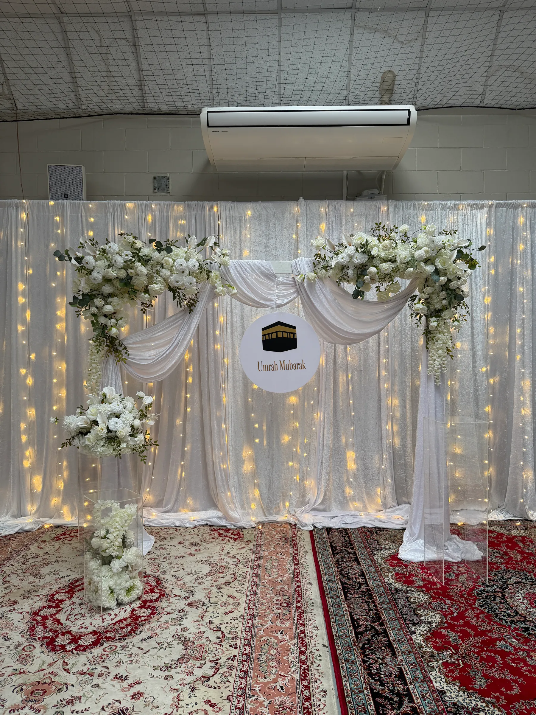 Cultural styling by Decoration X: White draped arch with white rose and hydrangea clusters and a round sign showing the Kaaba, fairy-lit white curtains behind and Persian rugs below.
