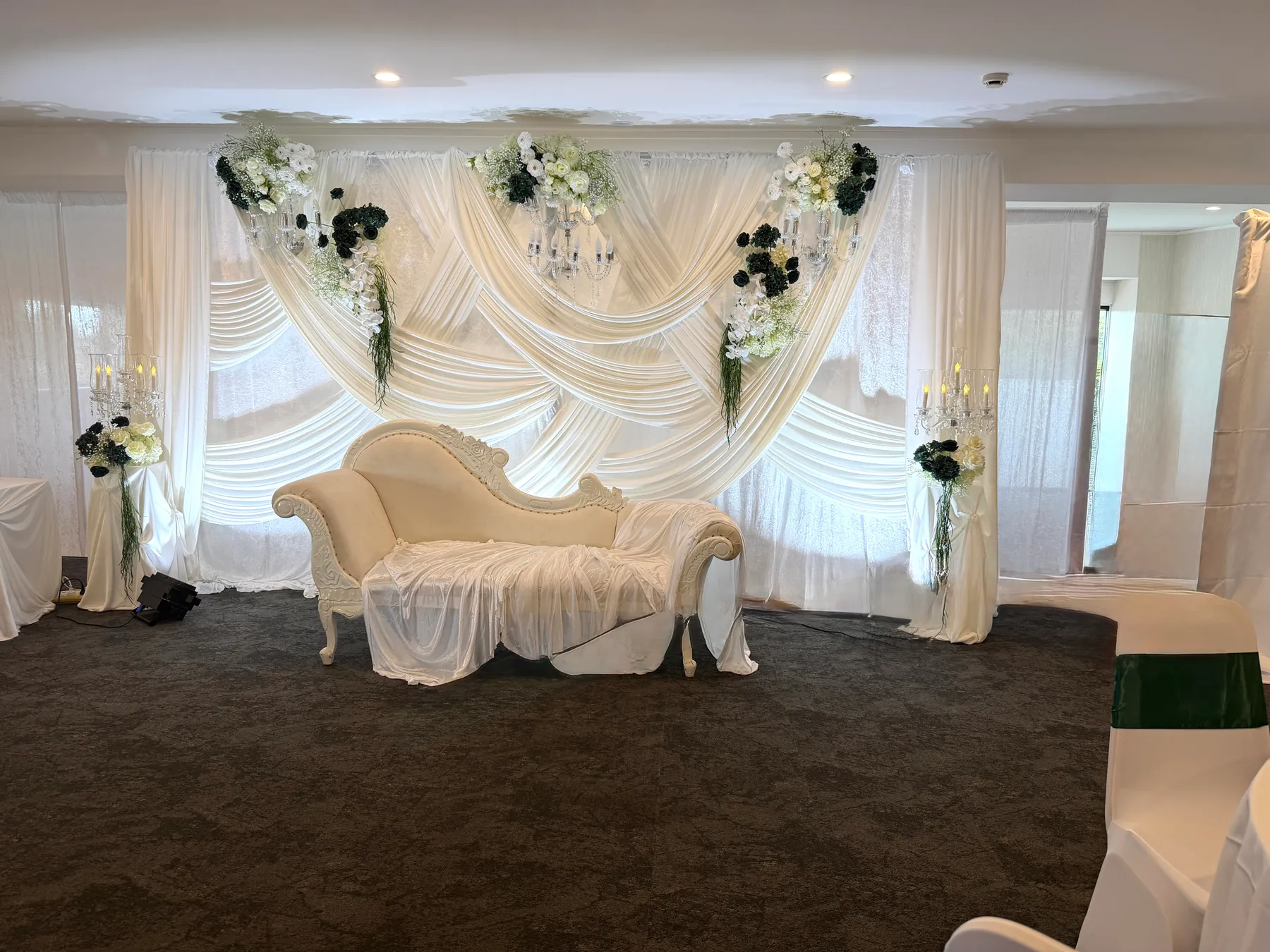 Stage styling by Decoration X: Ivory chaise draped in sheer fabric before crossed ivory swags with crystal candelabra and white and dark green florals, a sashed chair at the edge.