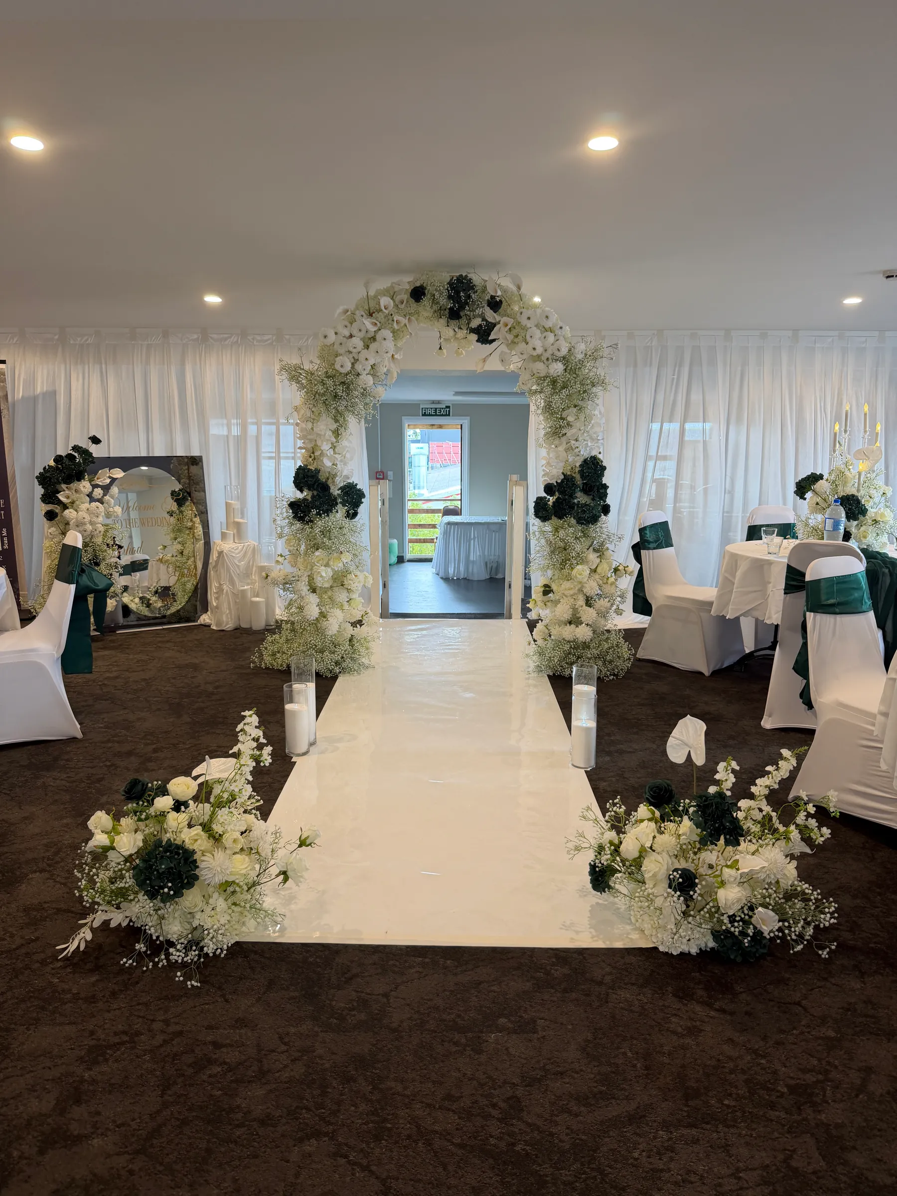 Ceremony styling by Decoration X: White aisle runner to a round arch of white roses, gypsophila and dark green roses, candles on the floor, sashed chairs and a mirror welcome sign to the left.