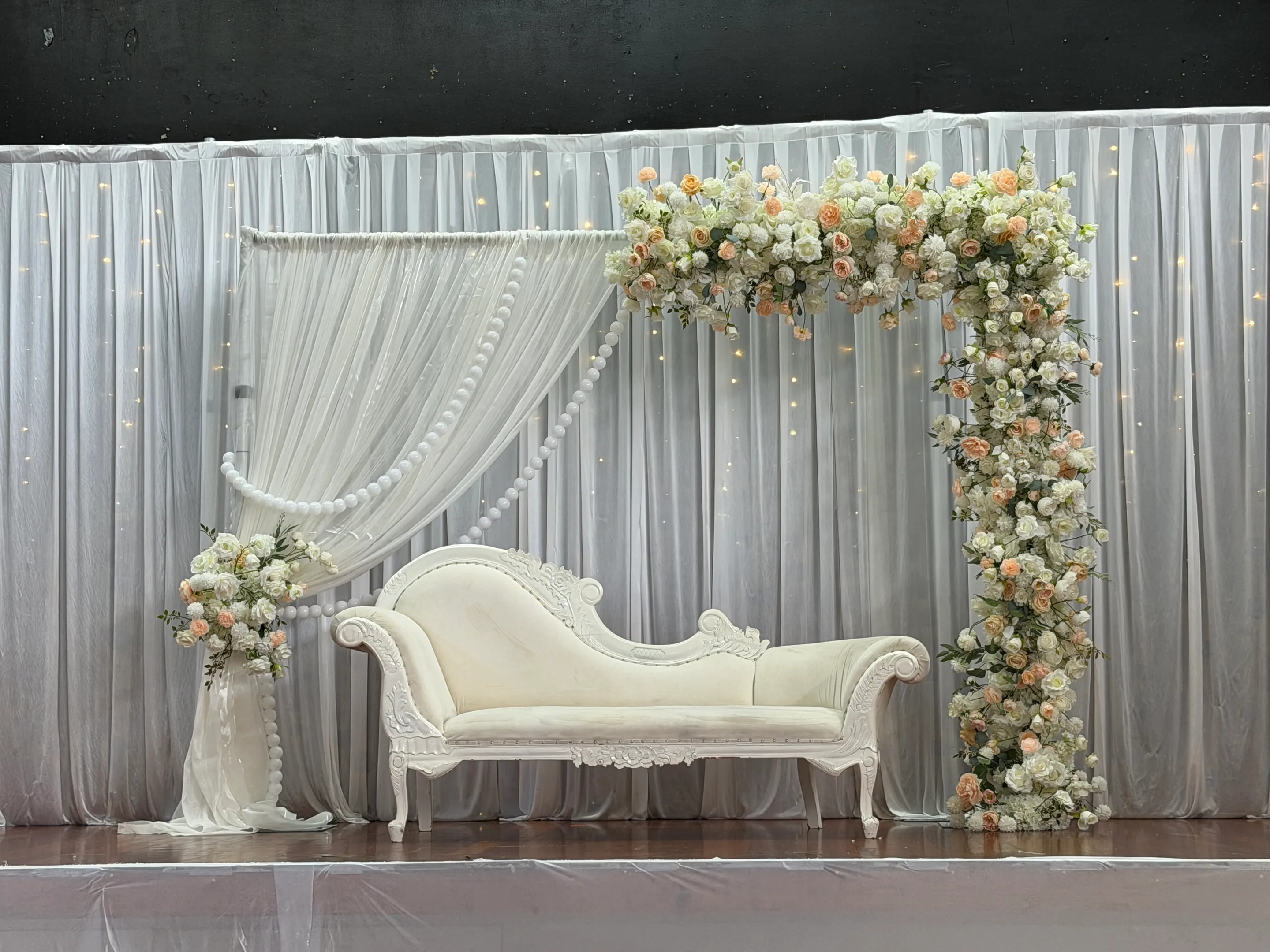 Stage styling by Decoration X: White carved chaise beneath an L-shaped garland of peach and ivory roses, a swagged sheer drape with pearl strands and fairy-lit white curtains behind.