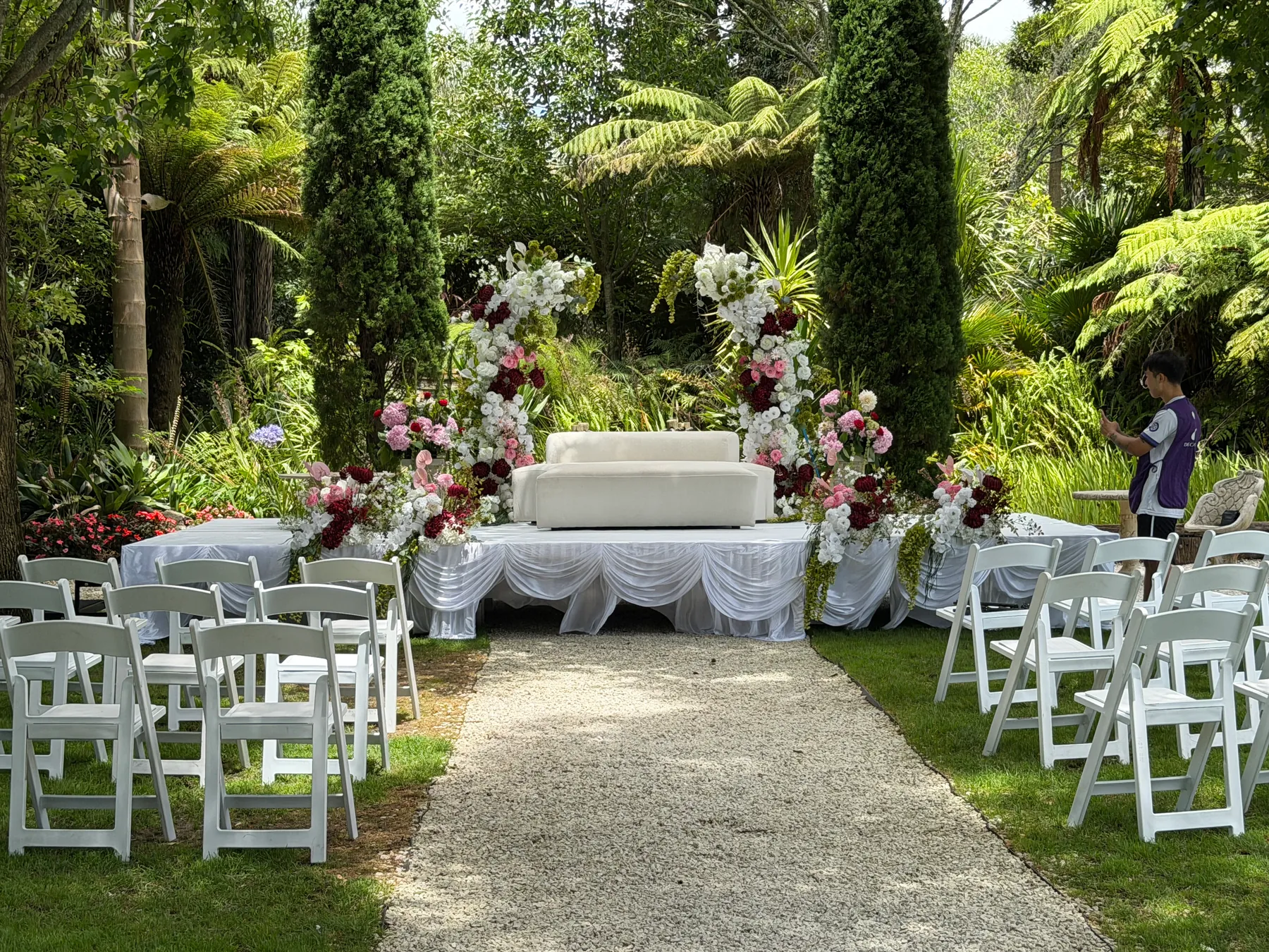 Ceremony styling by Decoration X: Gravel aisle between white folding chairs to a draped stage with a cream sofa and white, burgundy and pink floral columns, set among tree ferns and cypress.